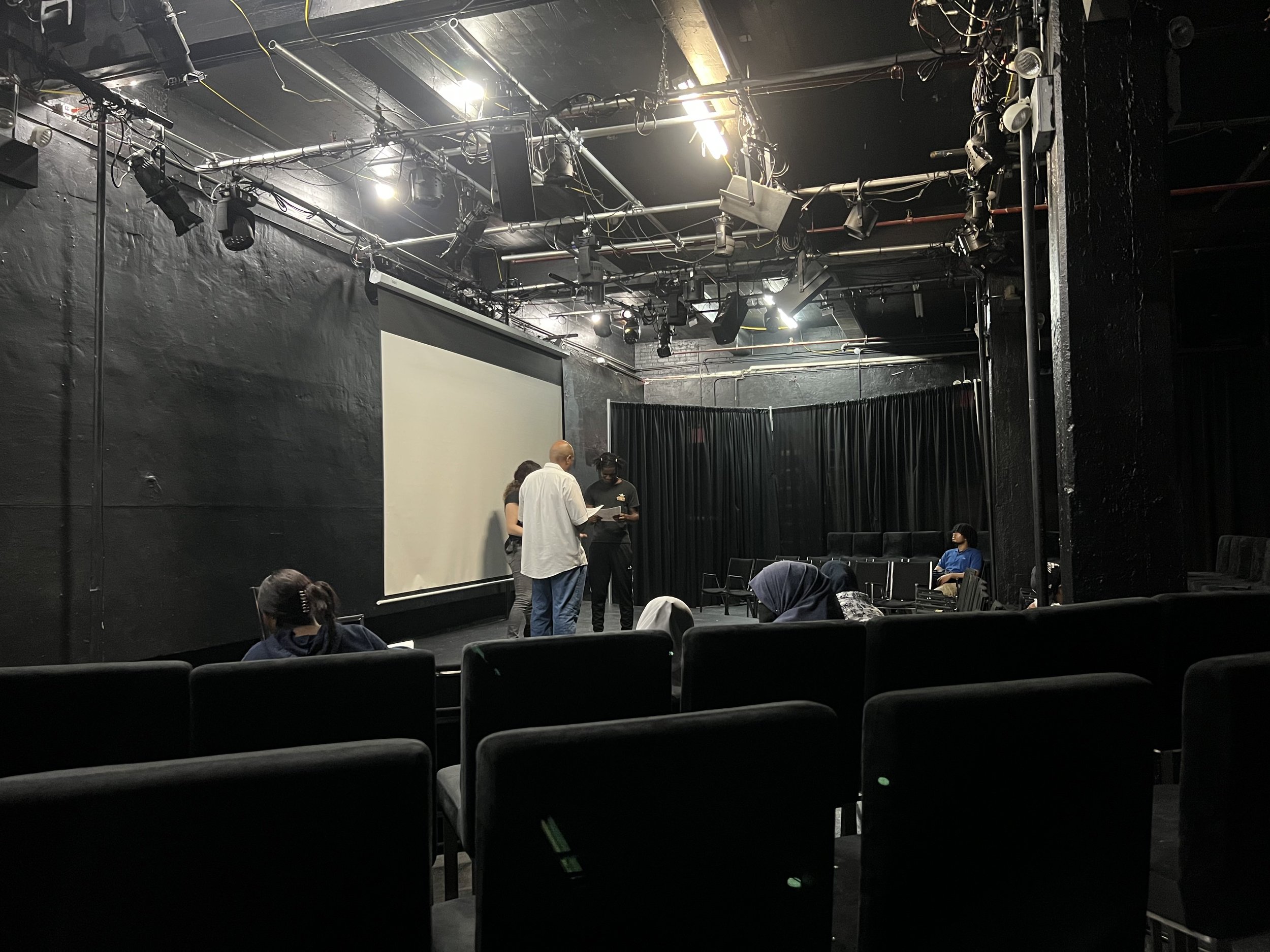 Empty theater with black walls, chairs, and a large white projection screen at the front. Several people are seen standing and sitting, preparing for a performance or presentation. Equipment and lighting are installed above the stage area.