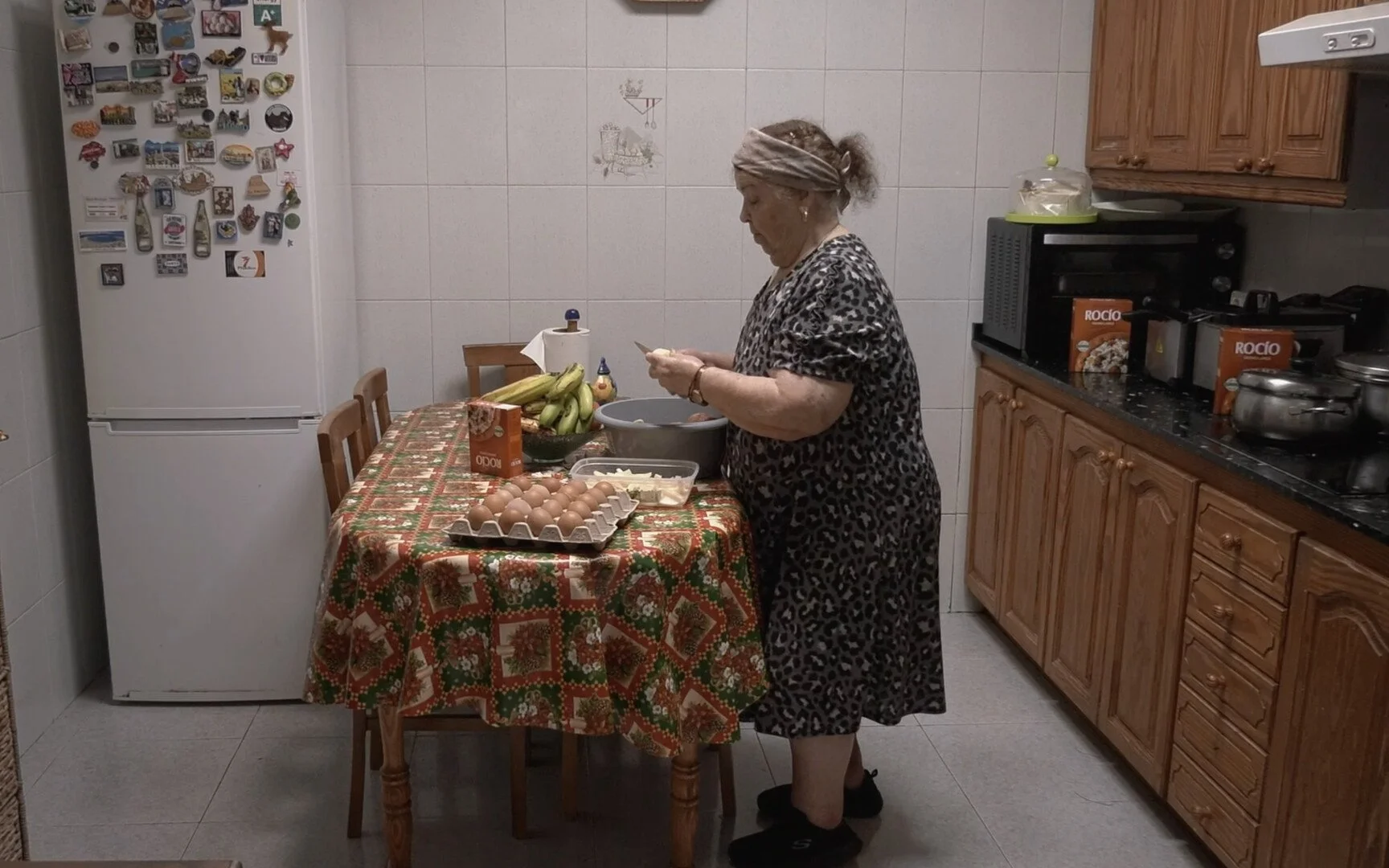 An elderly woman with a grey headscarf and a leopard print dress preparing food in a kitchen. She is standing at a table with a floral tablecloth, eggs, bananas, and food containers.