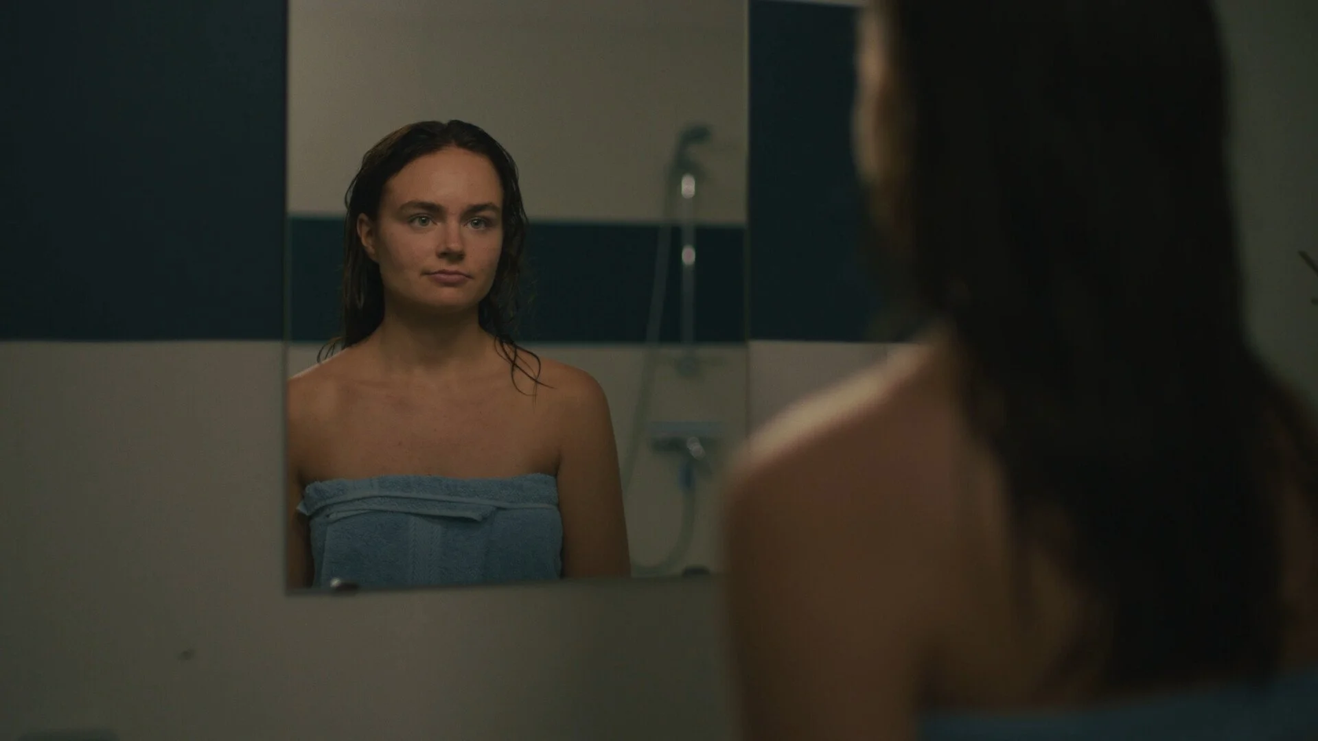 A woman with wet hair looking into a mirror in a bathroom, wrapped in a blue towel.