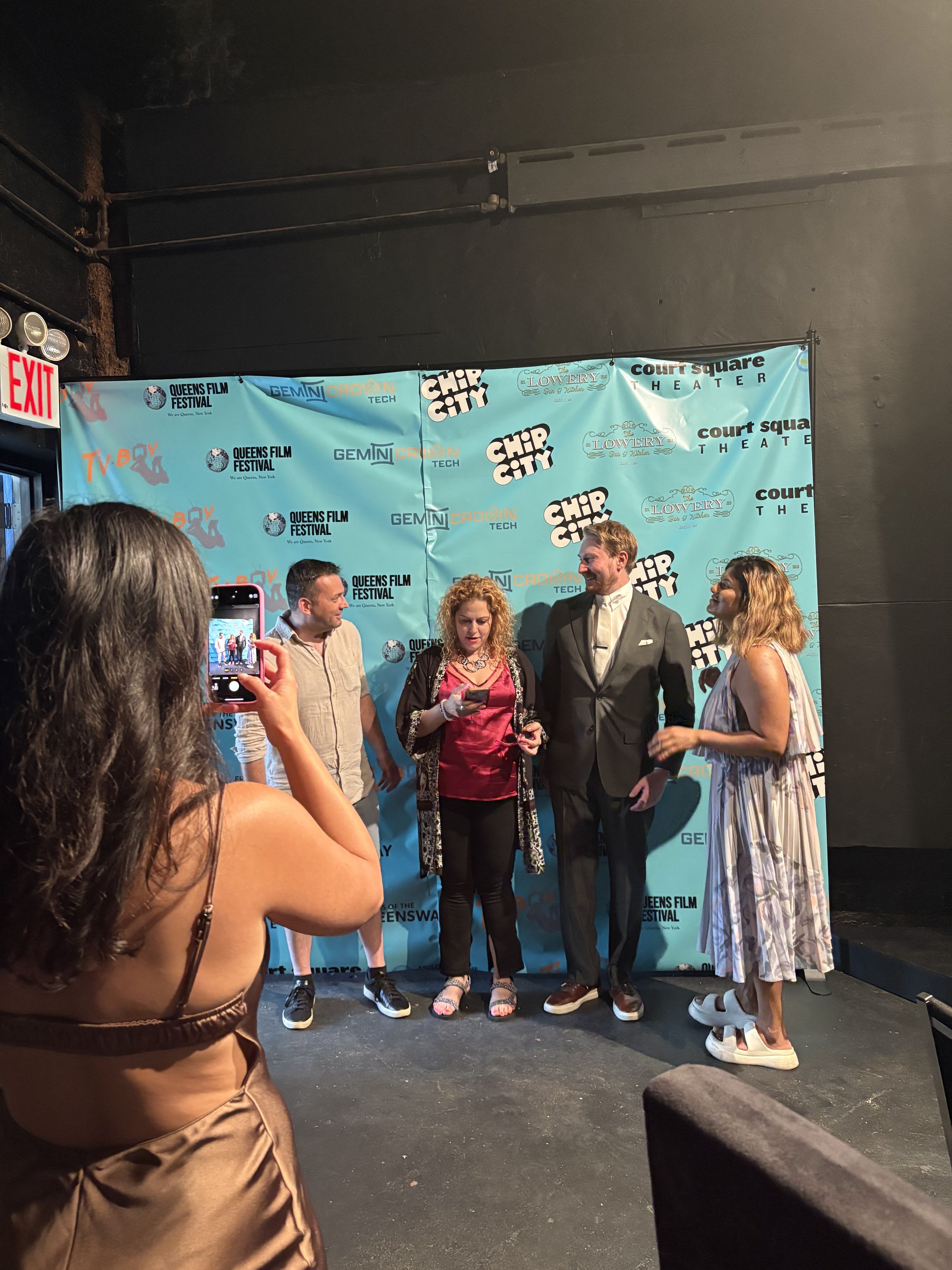 A woman in a bronze satin dress standing in front of a blue backdrop taking a photo with her phone of four people talking at an event.