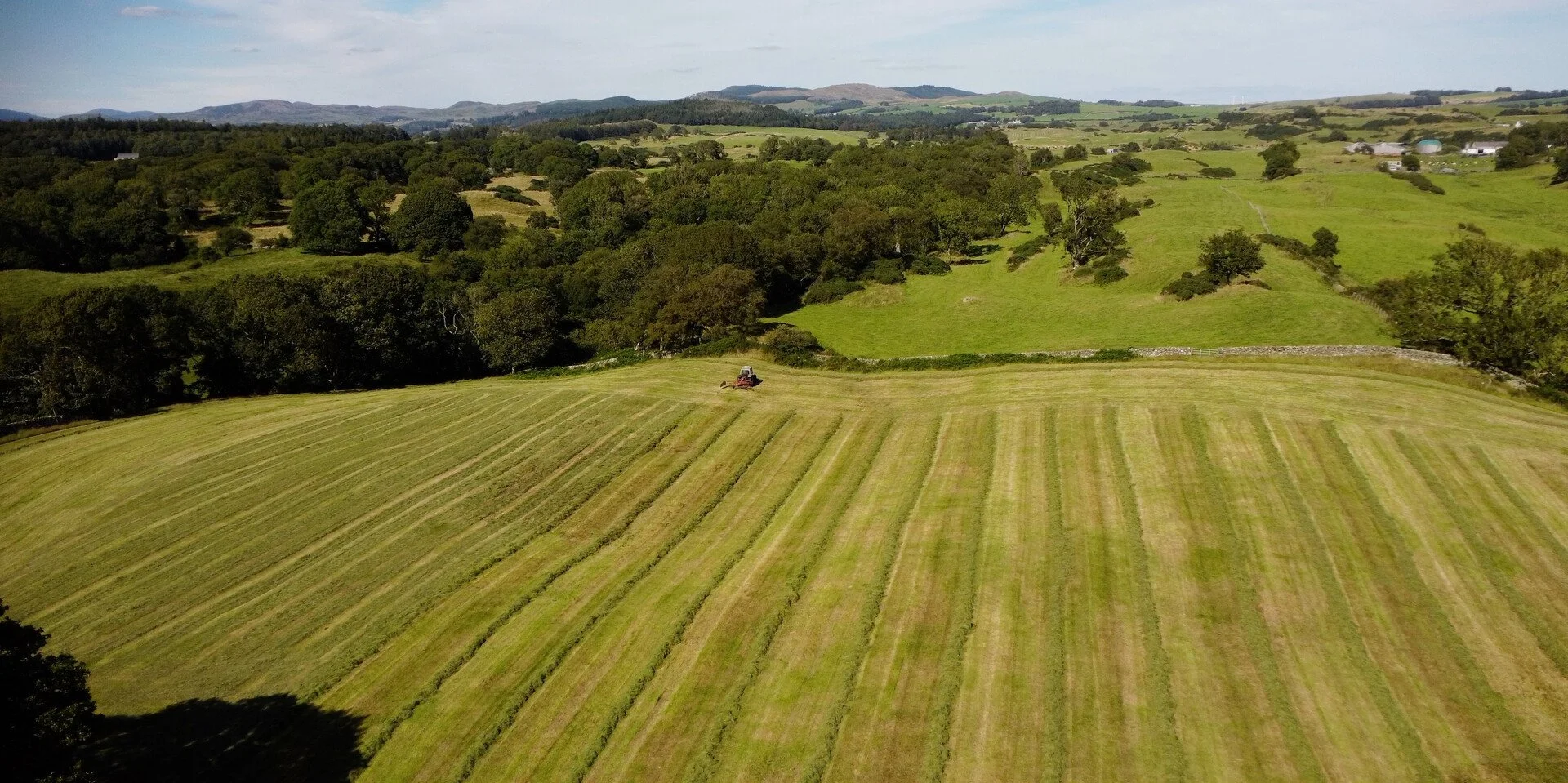 Aerial view of a lush green countryside with rolling hills, trees, and farm buildings in the distance. A tractor is working on freshly cut grass or hay in the foreground.
