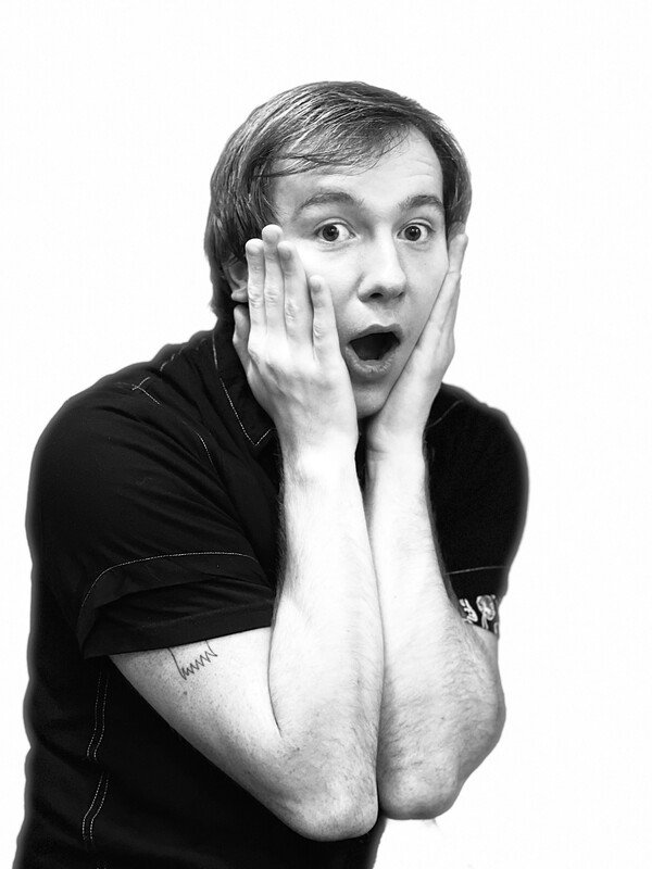Black and white photo of a surprised man with hands on his face, mouth open, wearing a short-sleeved black shirt.