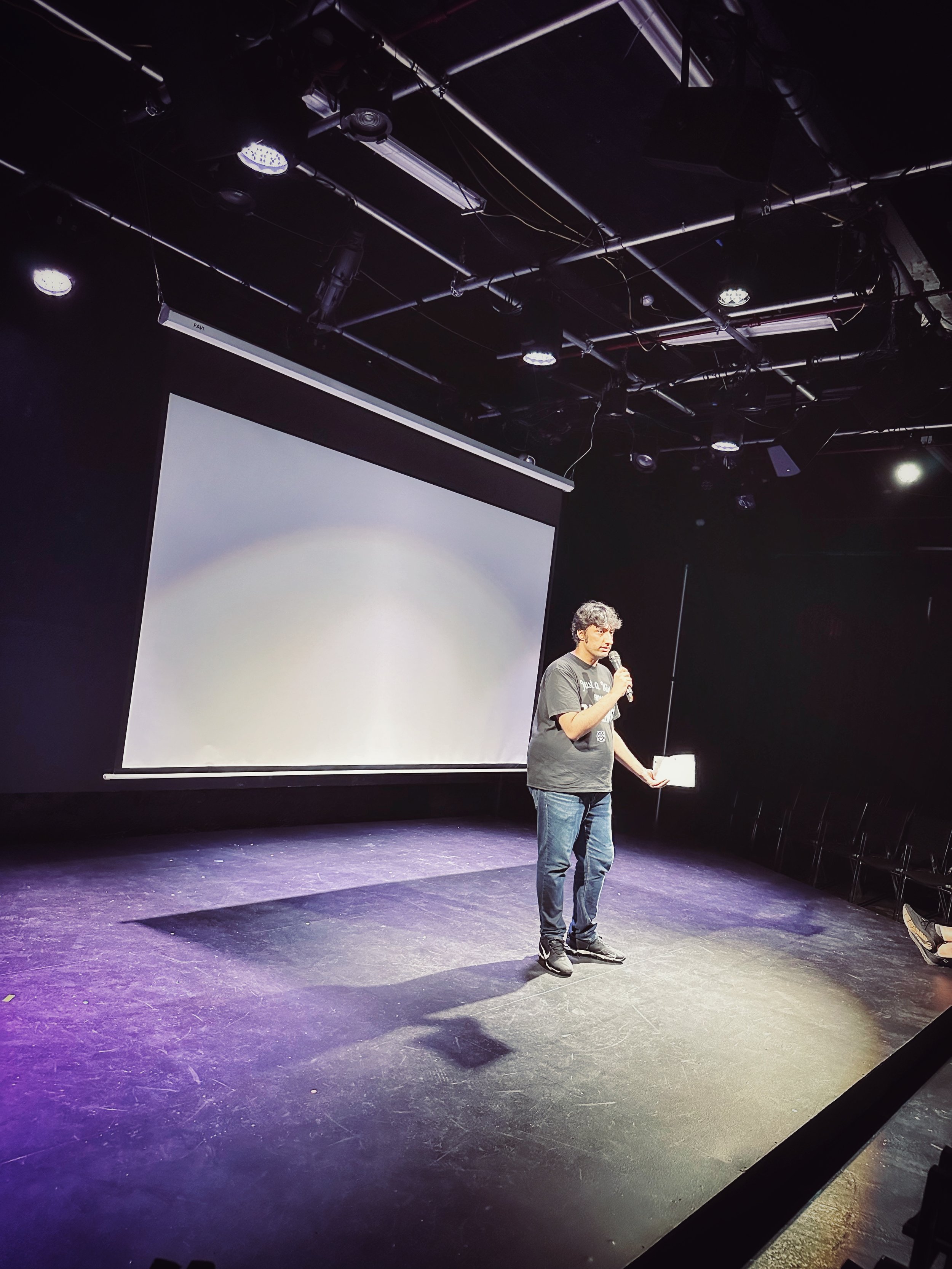A man standing on a stage with a microphone in one hand, in front of a large projection screen, in a dark theater or auditorium.