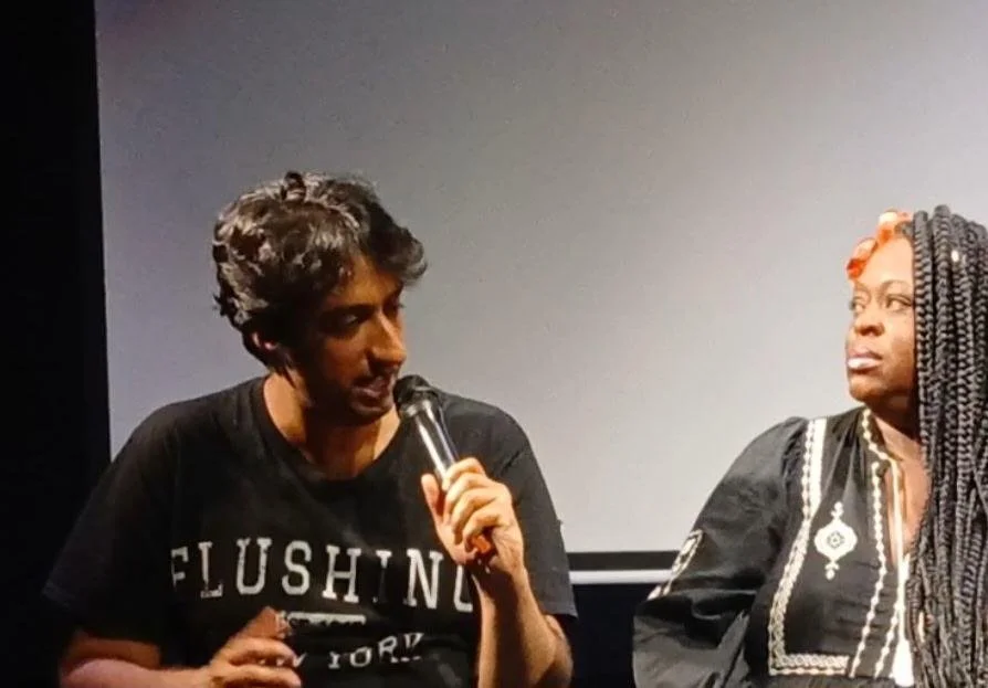 Two women engaged in a conversation, one holding a microphone. They are sitting in front of a plain backdrop, possibly at an event or panel.