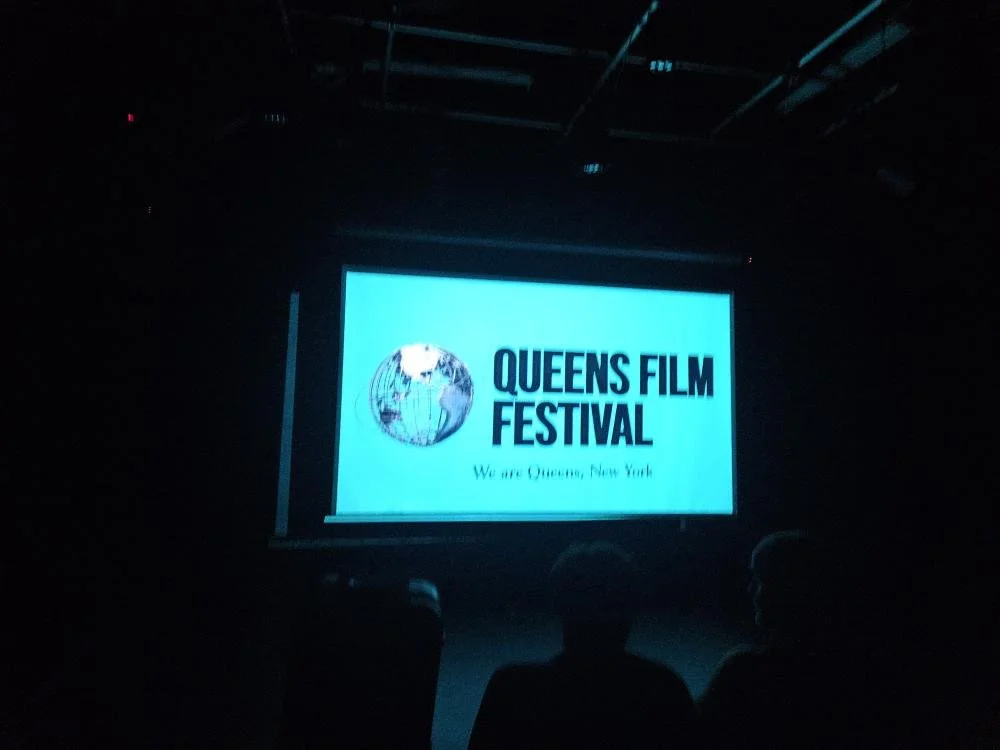Dark room showing a screen with the Queens Film Festival logo, featuring a globe and text, with silhouettes of audience members in front.