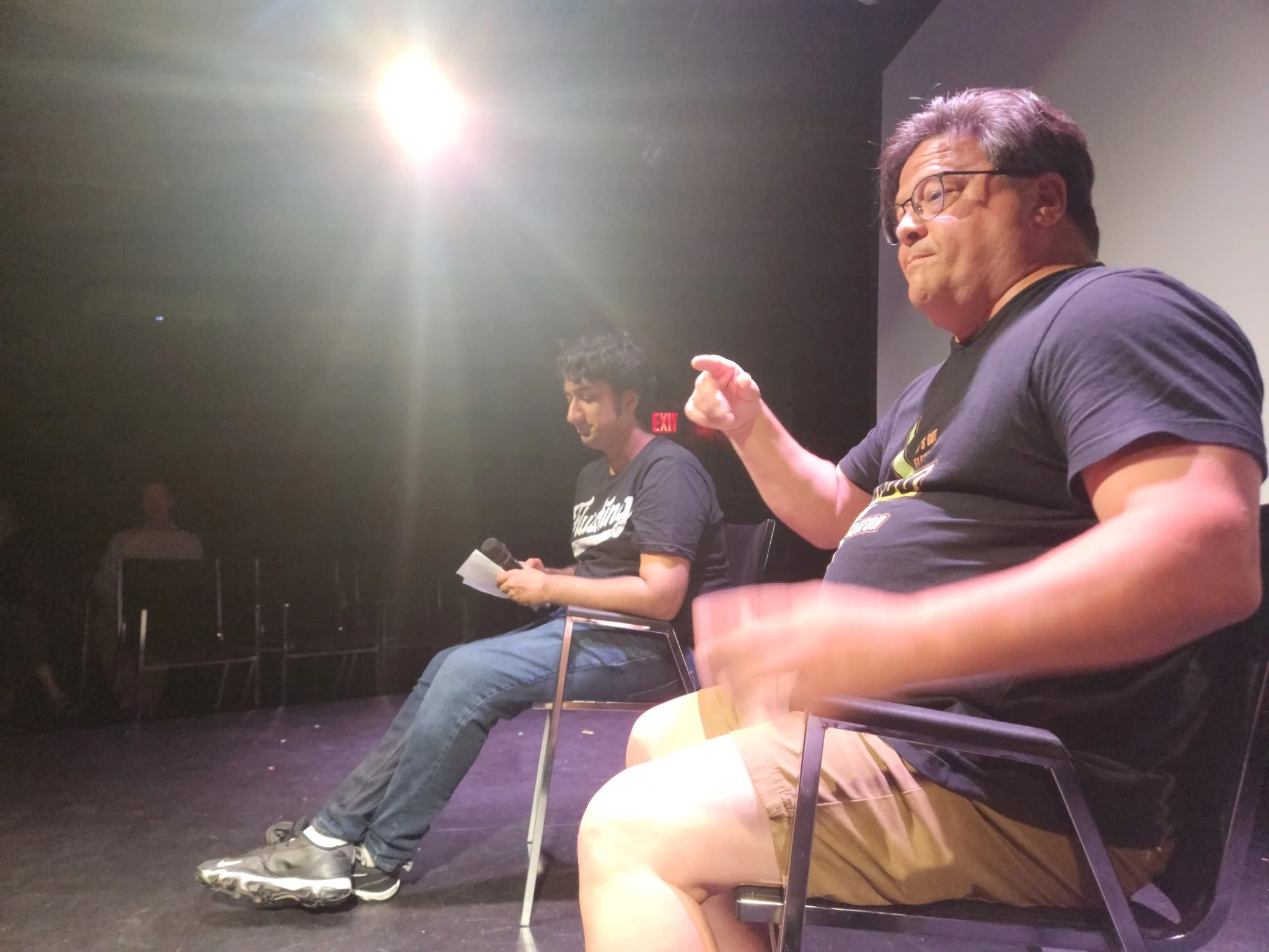 Two men are sitting on chairs on a stage, with one holding a microphone. The man on the right is wearing glasses, a black T-shirt, and khaki shorts, and is gesturing with his right hand. The man on the left is reading from a paper, wearing a black T-shirt, jeans, and sneakers. There is bright stage lighting overhead.