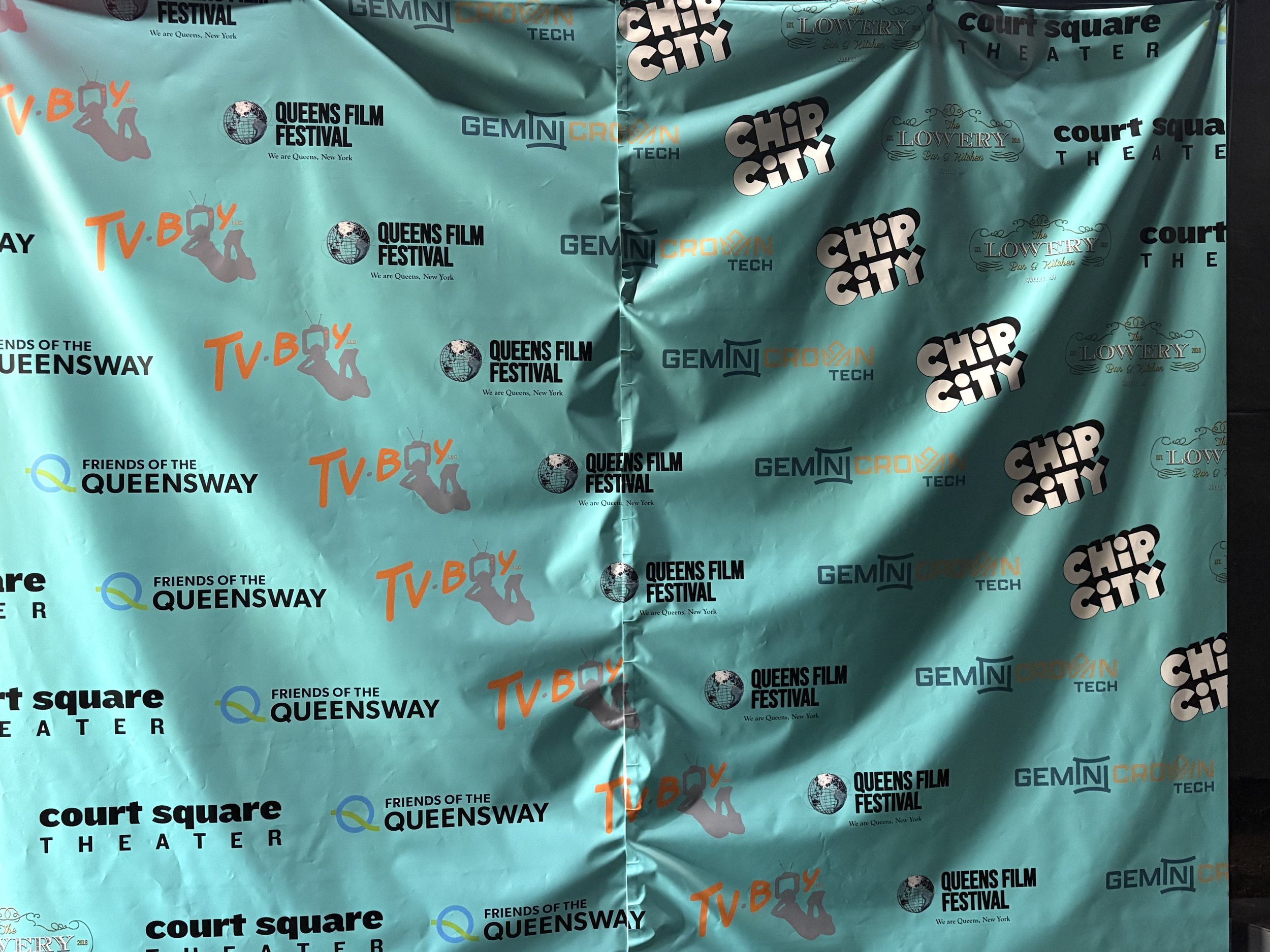 The backdrop features logos and names of several organizations and events, including Queens Film Festival, TV Boy, Friends of the Queensway, Chip City, Lowery Bus & Tiler, GEMINI Crown Tech, and Court Square Theater, set against a teal background.