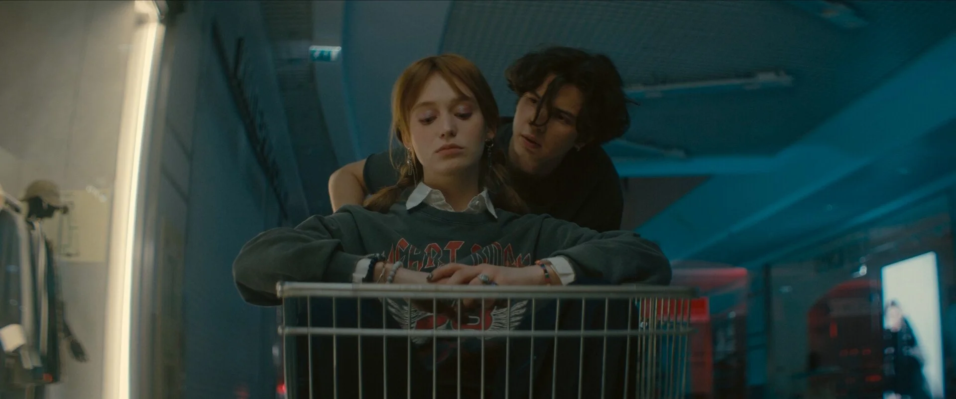 A young woman with red hair sitting in a shopping cart, looking down. A young man leaned over her, looking at her with concern. They are in a store aisle with shelves and a vending machine in the background.