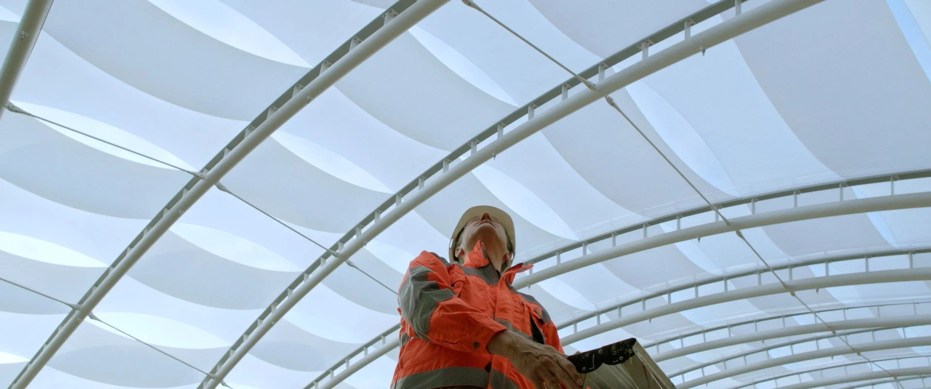 A person in an orange and gray jacket and wide-brimmed hat looking up at a modern architectural ceiling with white wavy panels.