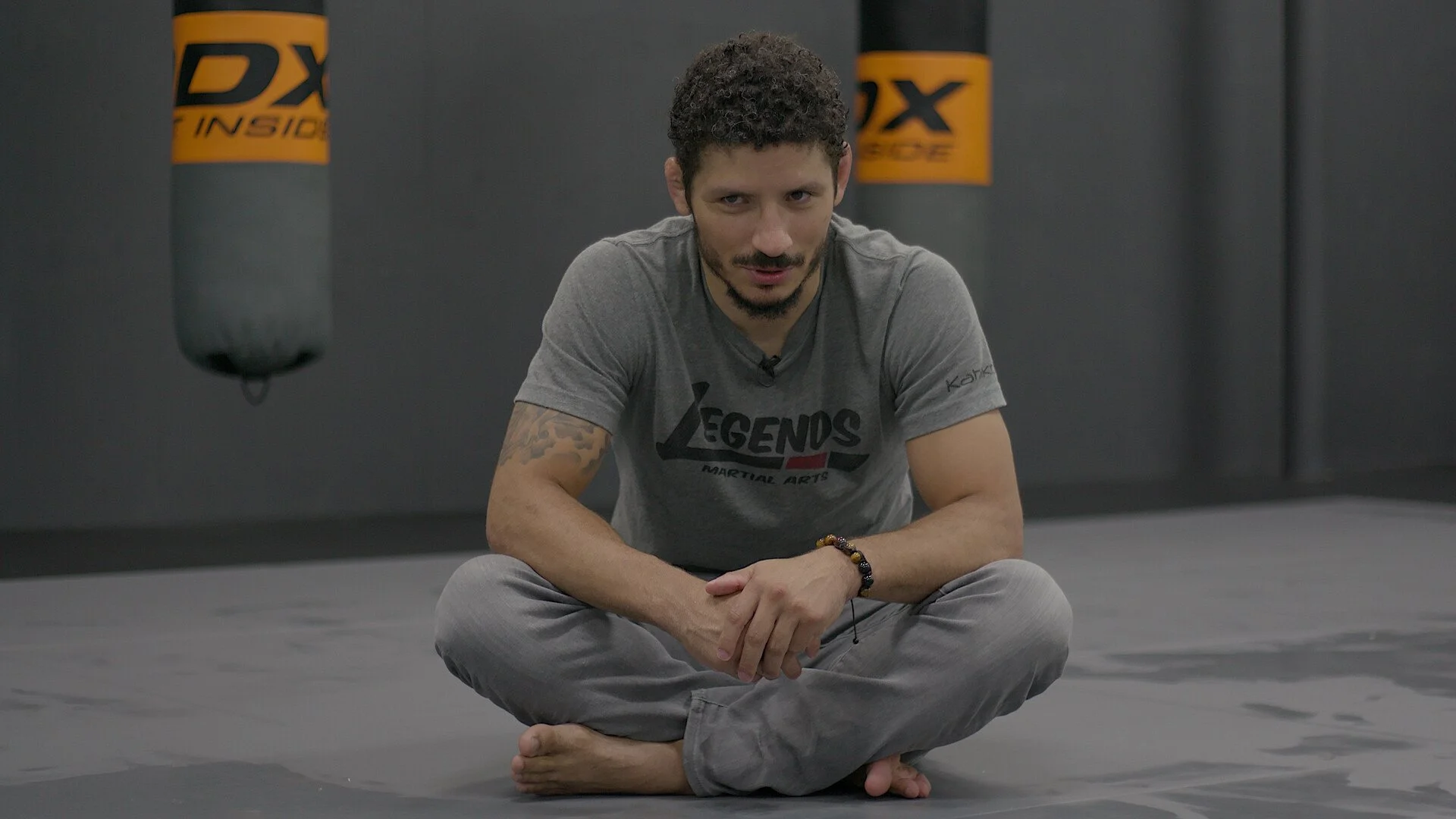 A man with curly hair and tattoos sitting cross-legged on the floor in a martial arts gym, wearing a grey t-shirt and pants, with punching bags hanging in the background.