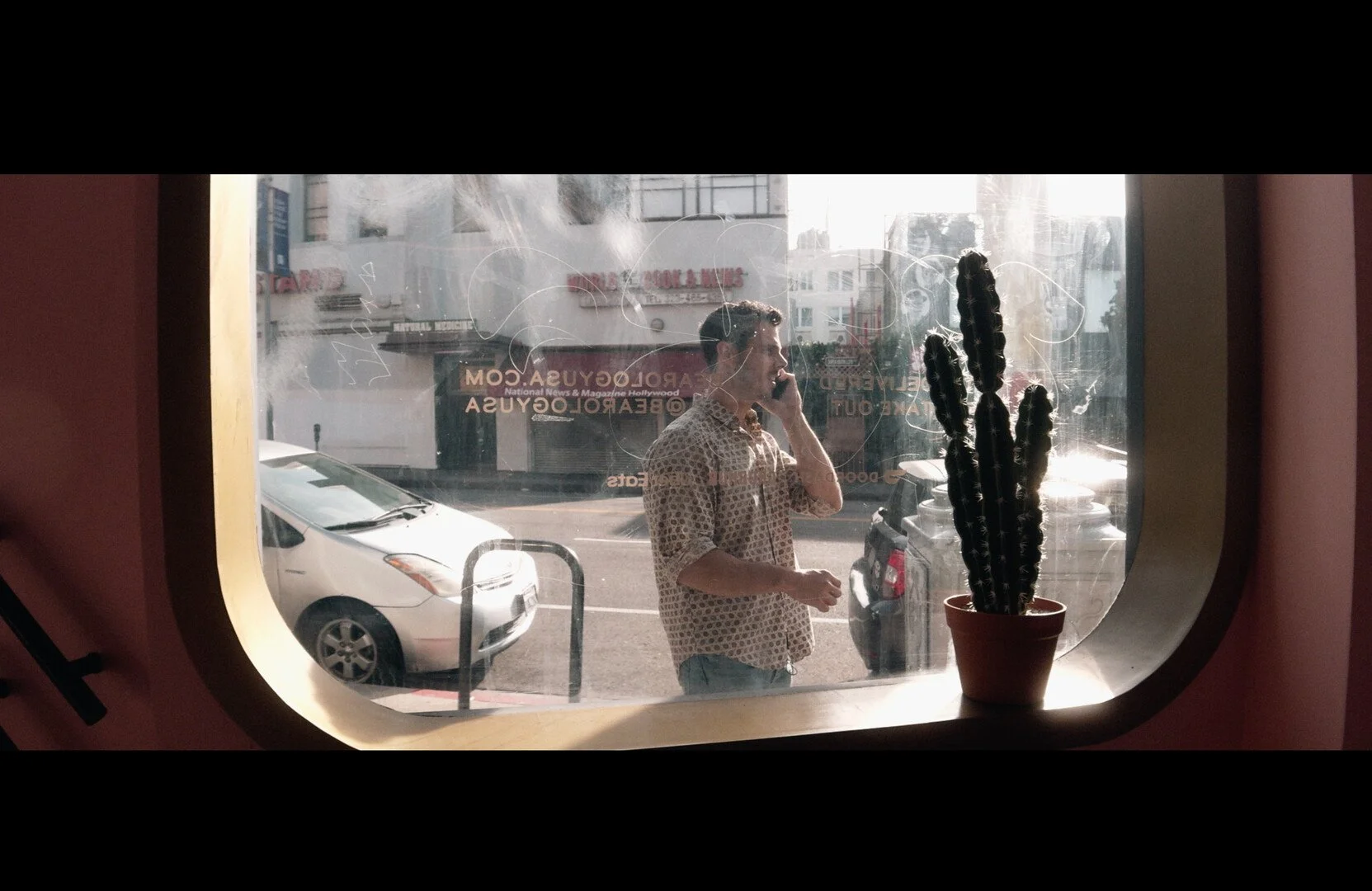 A man standing outside a window, talking on his cellphone. There is a potted cactus plant on the window sill, and parked cars on the street beyond.