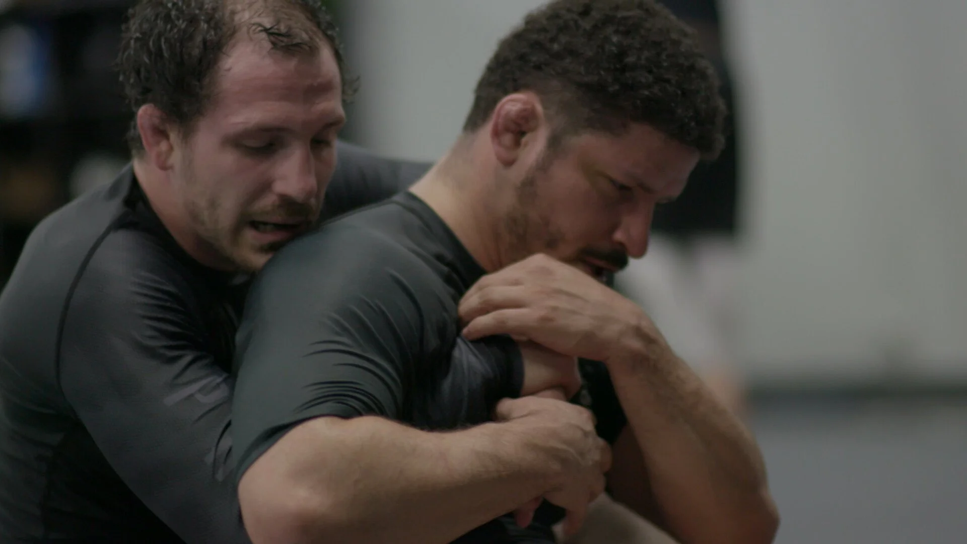 Two men engaged in a physical fight, one man in a black shirt grabbing the other man from behind in what appears to be a martial arts or self-defense training session.