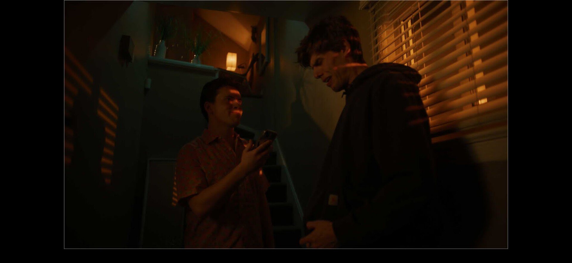 Two young men standing near a staircase in a dimly lit room with blinds on the window. One man is looking at his phone with a slight smile, while the other man is smiling and looking at him. There is a warm light source and a small lamp in the background.