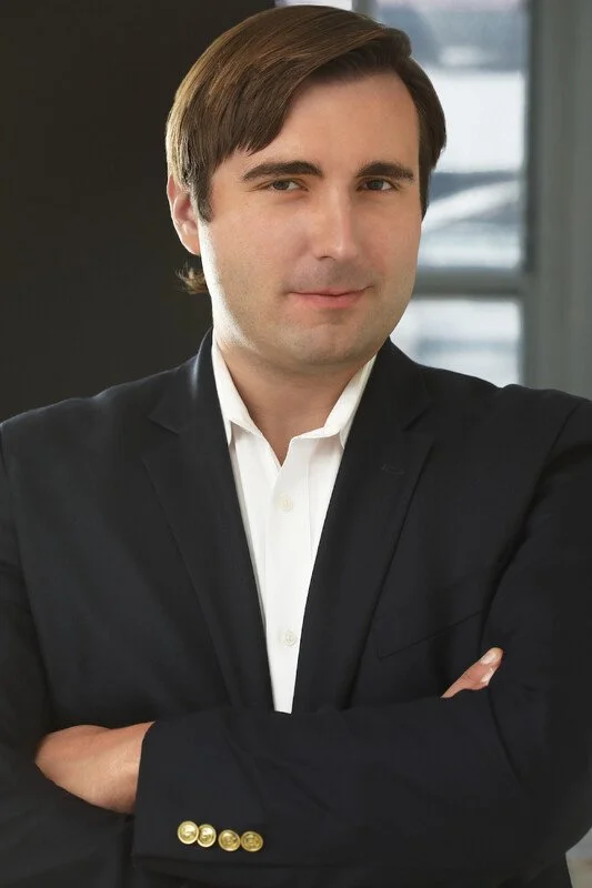 A man in a dark suit with a white shirt, standing with arms crossed in an office setting with windows in the background.