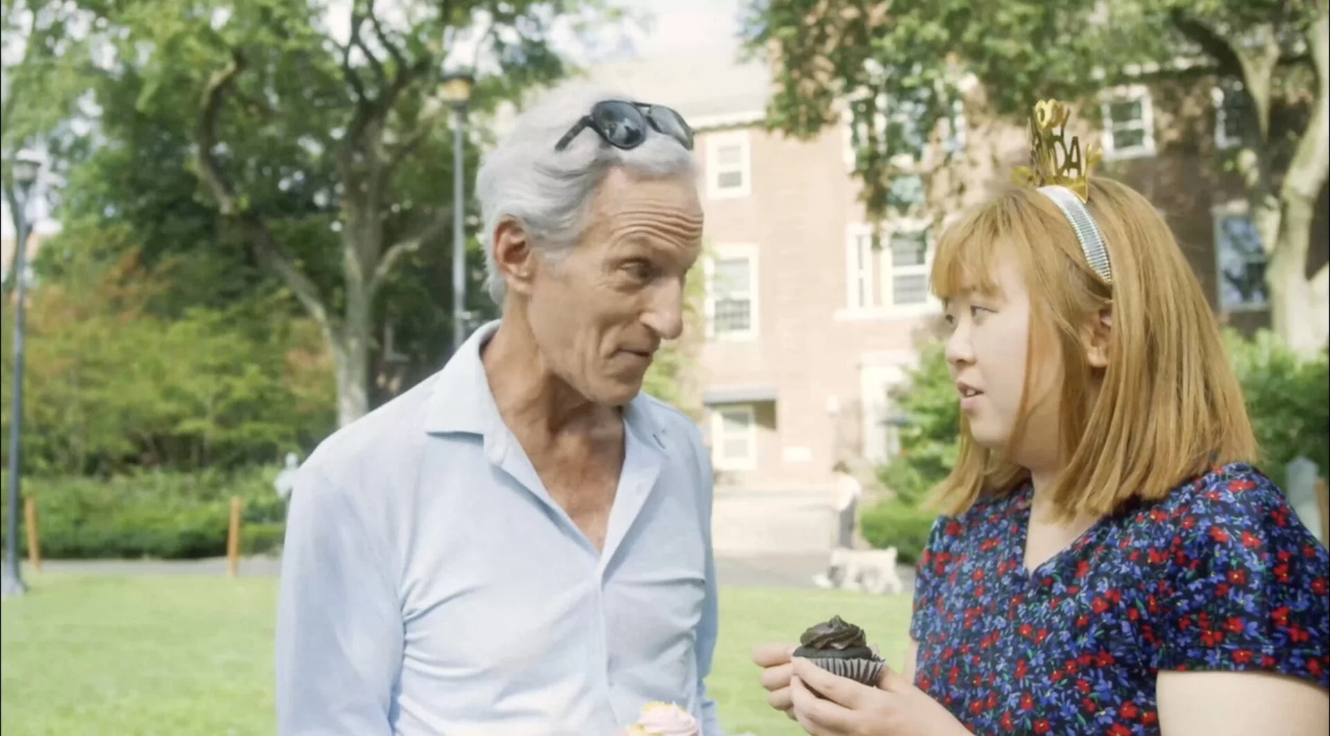 A young girl with red hair wearing a birthday tiara and a floral dress holds a chocolate cupcake, talking to an older man with white hair and sunglasses on his head, in a park with green trees and a brick building in the background.
