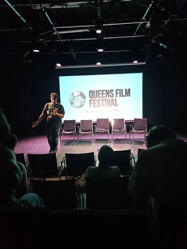 A person stands on stage in front of a screen that reads 'Queens Film Festival'. The stage has several empty chairs and audience members seated facing the stage.