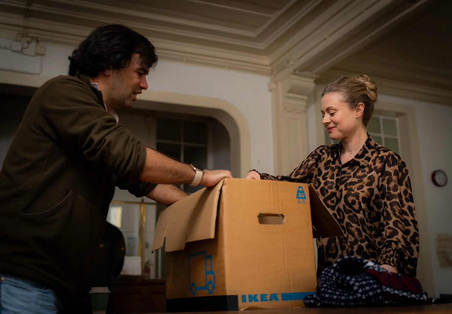 A man and woman are packing or unpacking a cardboard IKEA box. The woman is smiling and wearing a leopard print top, while the man is leaning over the box, smiling. They are indoors in a warmly lit room.