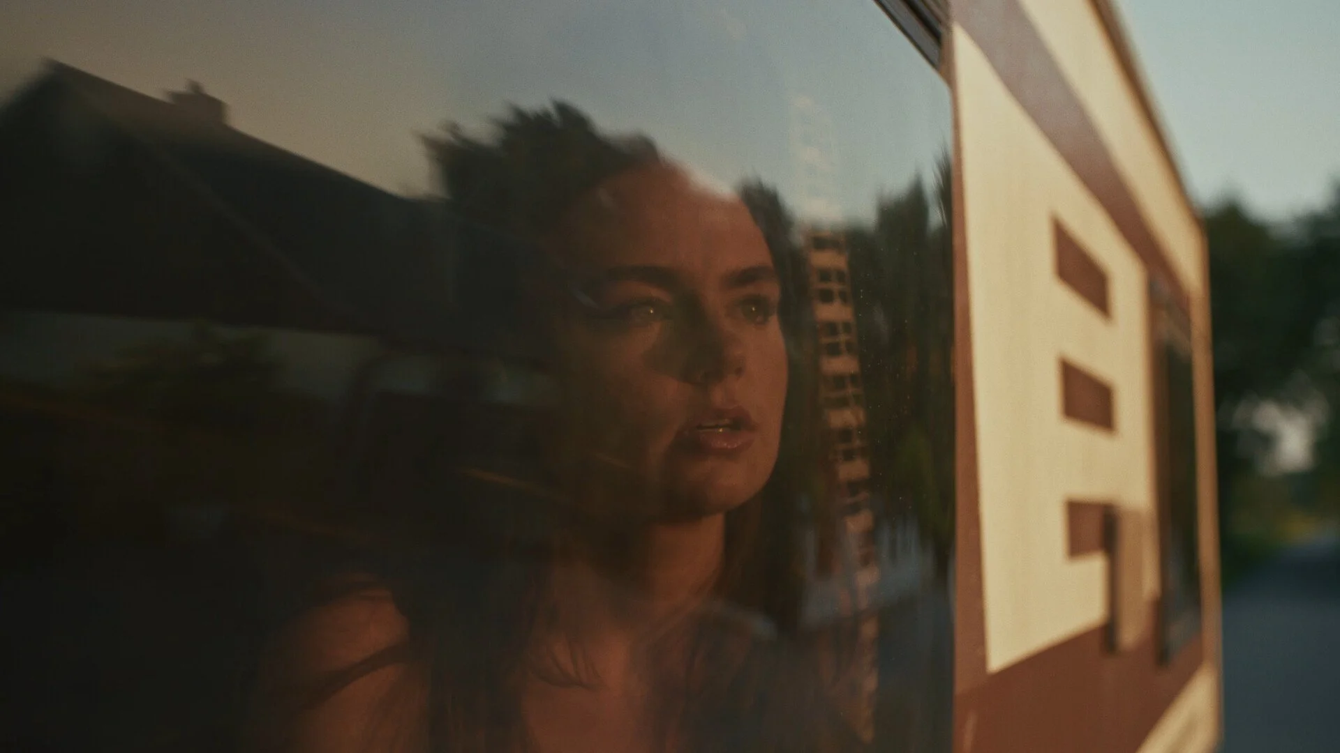 A woman looks out a bus window as the evening sun sets behind her, creating a reflection on the glass.