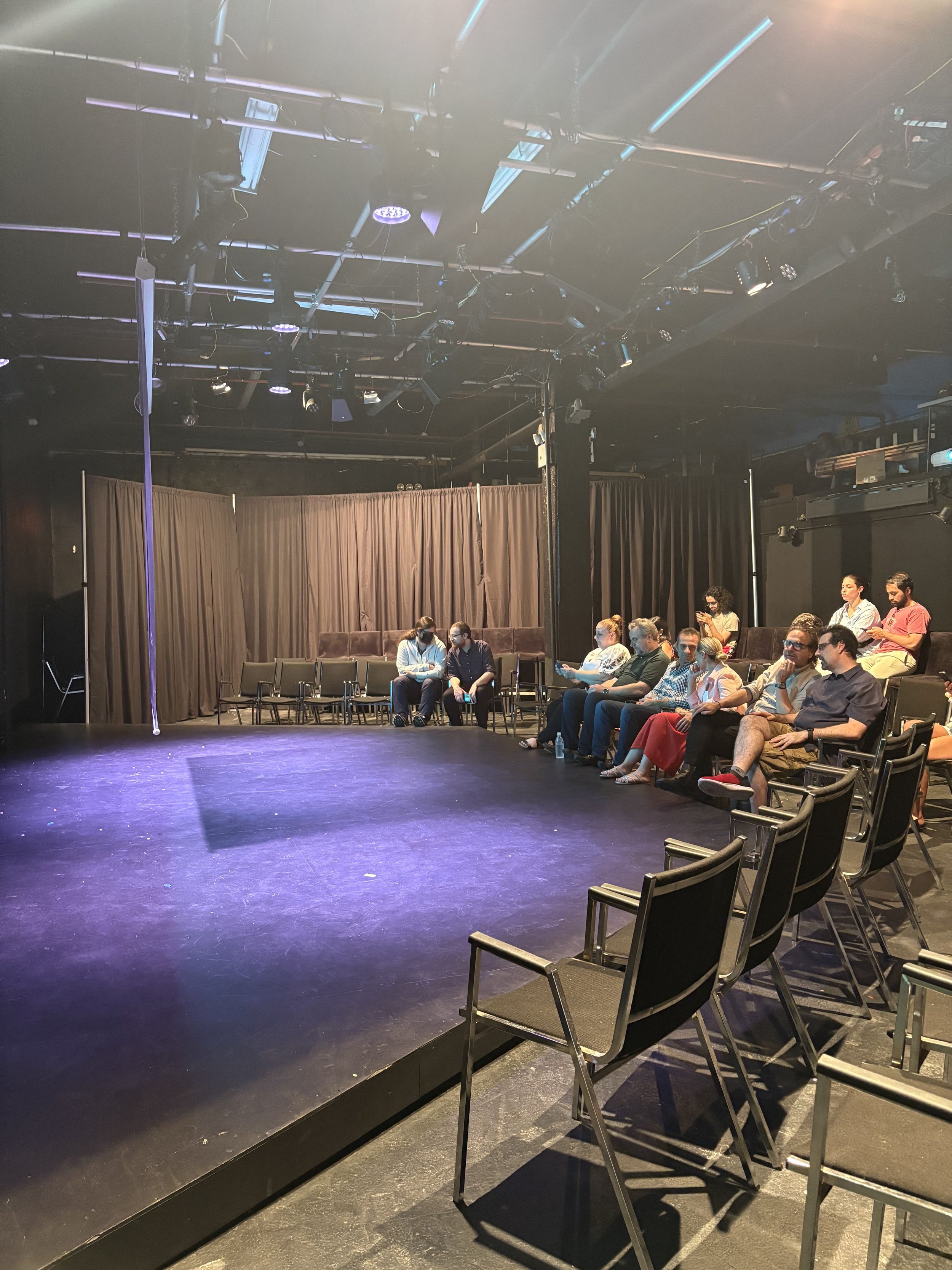 An empty theater stage with a few audience members seated on the right side, backstage black curtains, stage lighting equipment overhead, and chairs in the audience area.