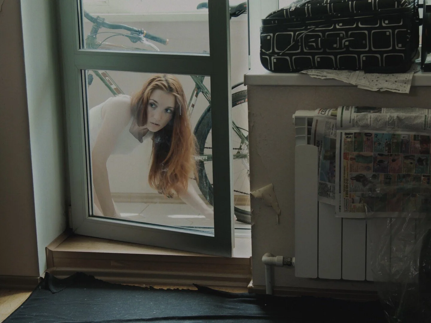 A woman with long red hair looking through a glass door from inside a room. Behind the door, a bicycle is visible. Inside the room, there are various items including newspapers, a black patterned bag, and a radiator.