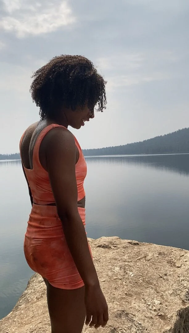 A woman with dark, curly hair stands on a rock by a calm lake, wearing a matching orange athletic top and shorts, looking down with a peaceful expression.