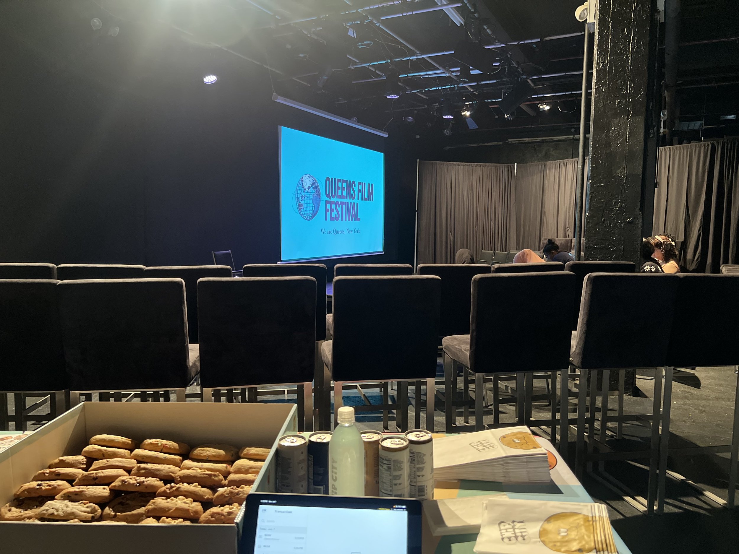 Empty theater with rows of black chairs in front of a large screen displaying the Queens Film Festival logo, cookies, drinks, and informational pamphlets on a table in the foreground, and a few people sitting at the back.