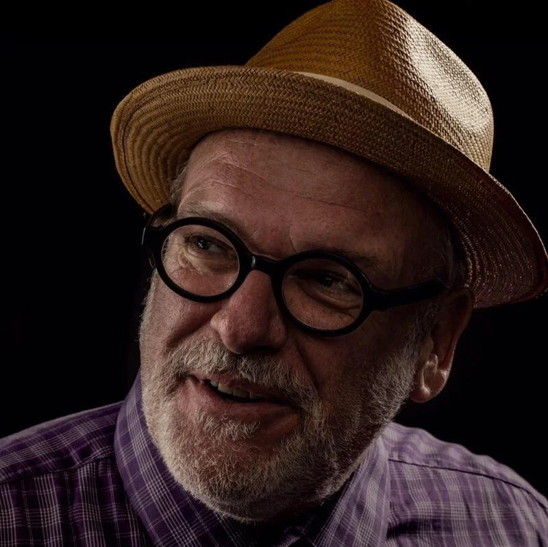 A man with a beard and glasses wearing a straw hat and purple plaid shirt, smiling against a dark background.