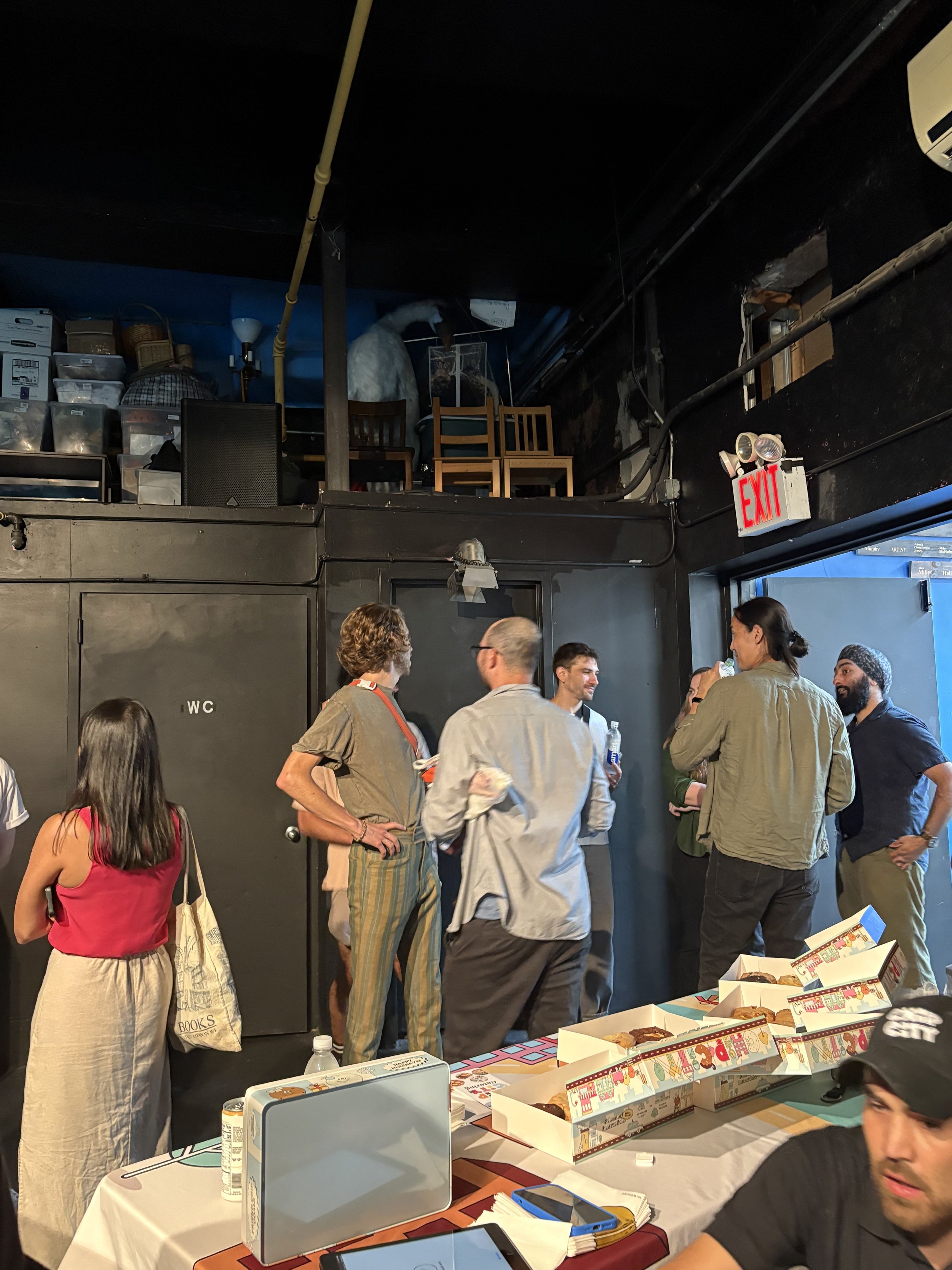 A group of people standing and talking in a room with a black wall, some chairs on an upper level, and boxes of donuts on a table in the foreground.
