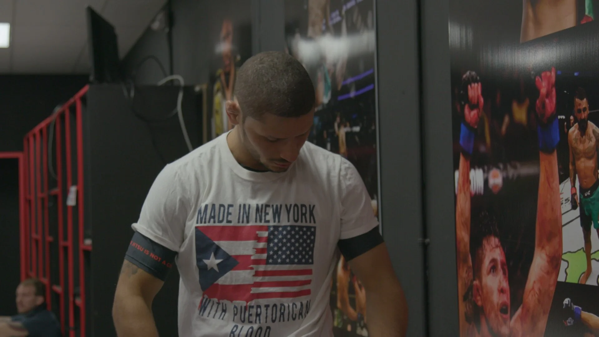 A man with short dark hair wearing a white T-shirt with a Puerto Rican flag design and text, standing near a wall with boxing posters, in a gym or training facility.