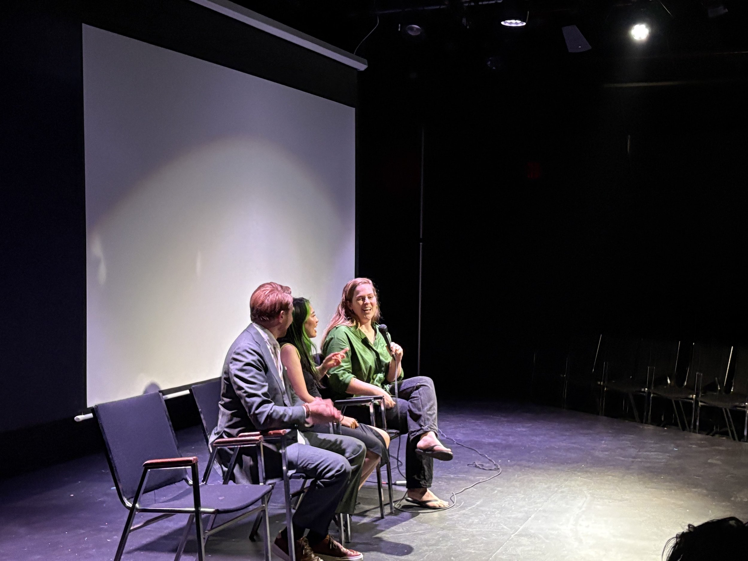 Three people sitting on chairs on a stage, engaged in a conversation during a panel discussion or interview. A woman holds a microphone and appears to be speaking with a smile. The stage has a large white screen or backdrop behind them, and the audience area is dark.