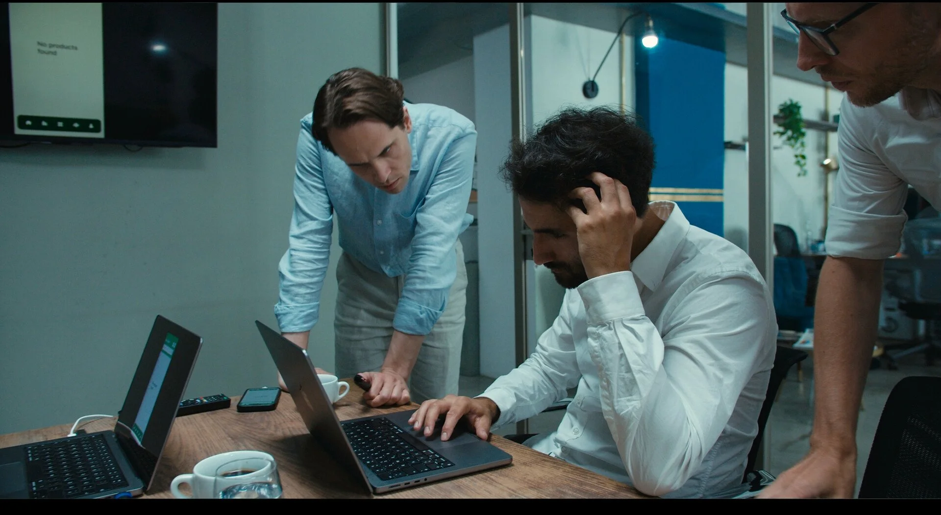 Three men in a meeting look concerned as they work on laptops around a table in an office.