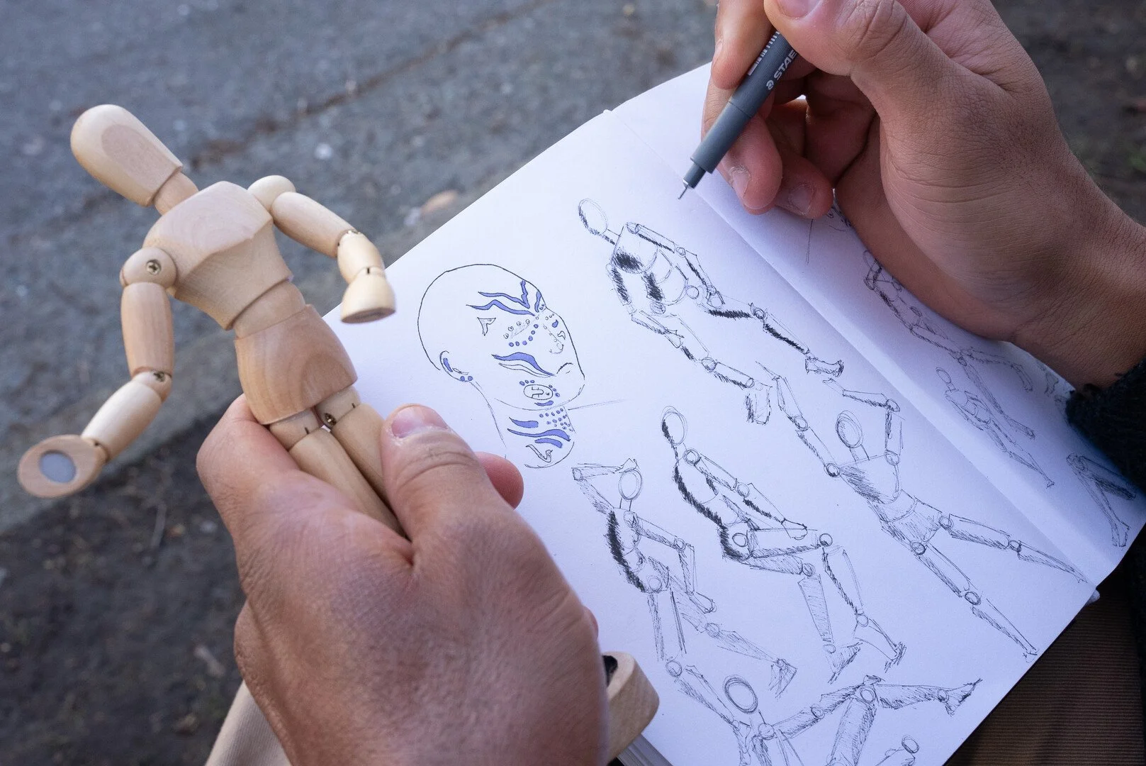 A person drawing a humanoid robot and anatomical sketches of a human head and body on paper, holding a small wooden mannequin in their left hand.