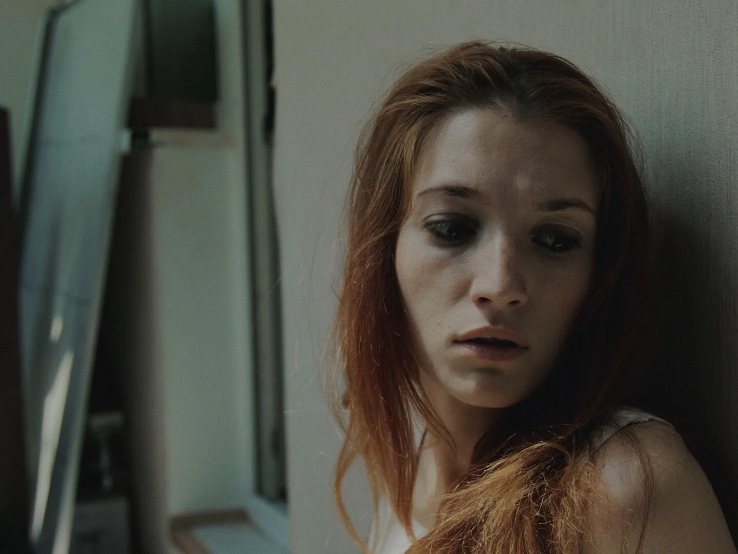 A woman with red hair and dark eye makeup leaning against a wall, looking down with a somber expression.