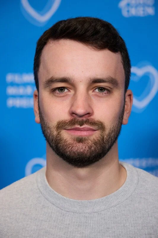 A young man with short dark hair and a beard, wearing a gray crew neck shirt, standing in front of a blue background with white heart icons and text.