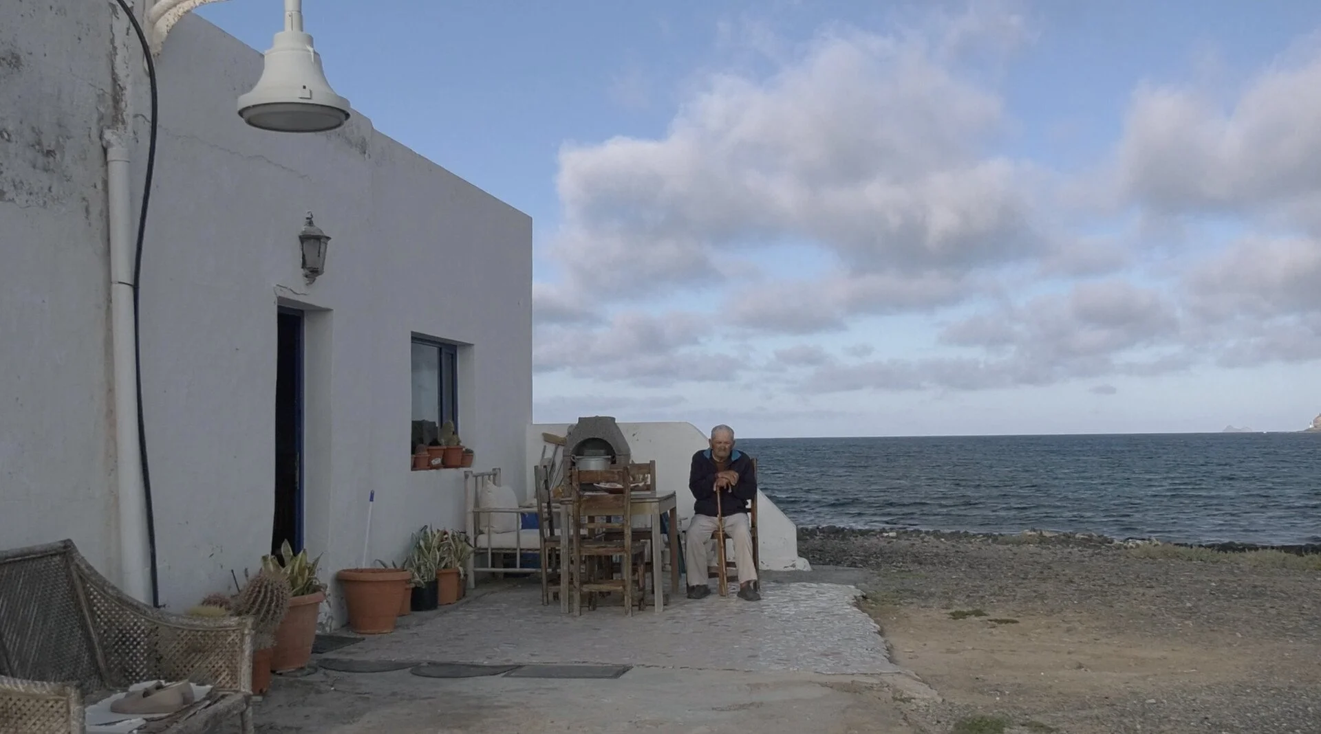 An elderly man sits on a wooden chair outside a white building near the ocean, with a patio table and potted plants, surrounded by cloudy sky and rocky shoreline.
