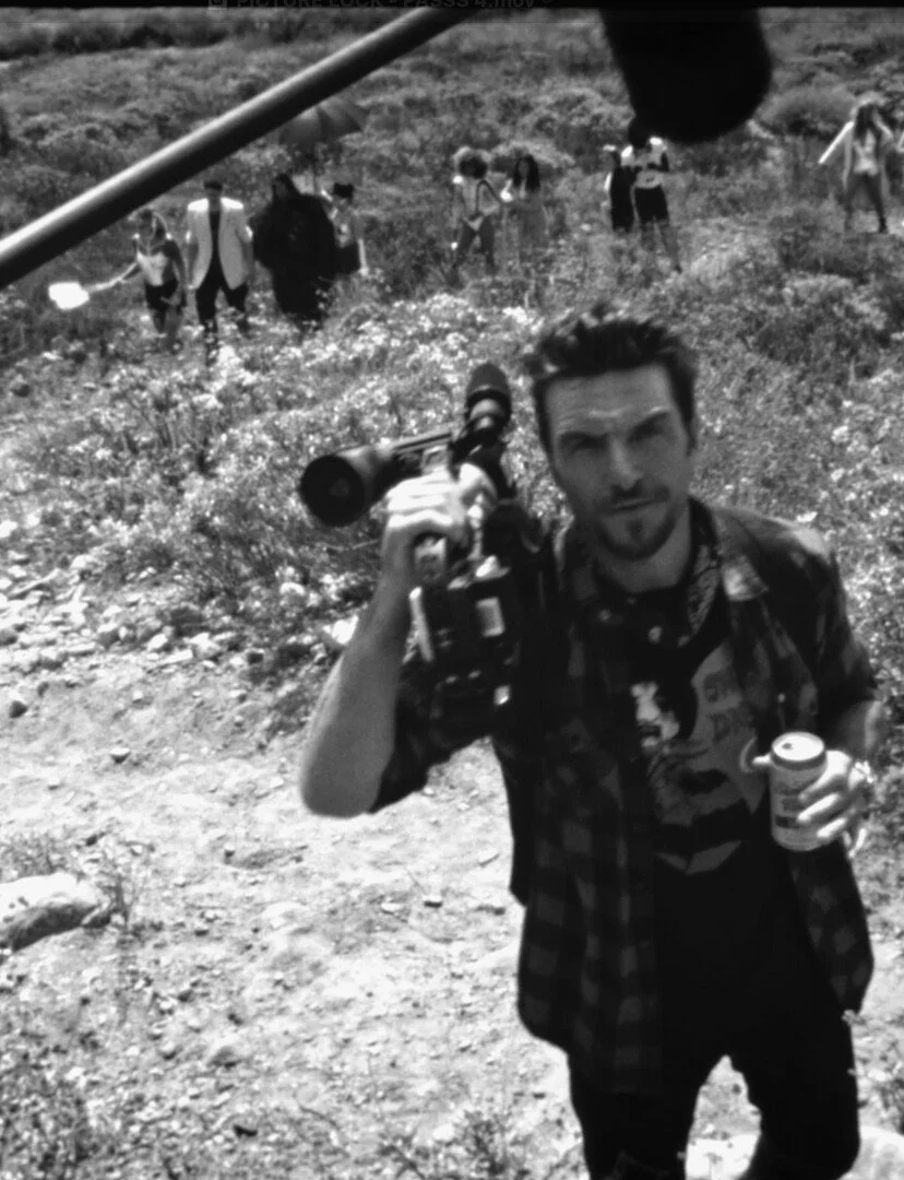 Photograph of a man holding a camera and a soda can in a dirt outdoor setting with other people in the background.
