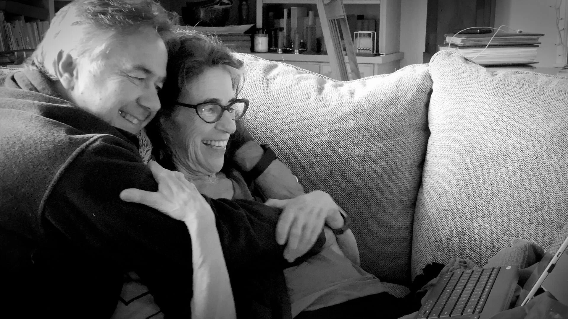 A man and woman sitting on a couch, smiling and hugging, while looking at a tablet or laptop.