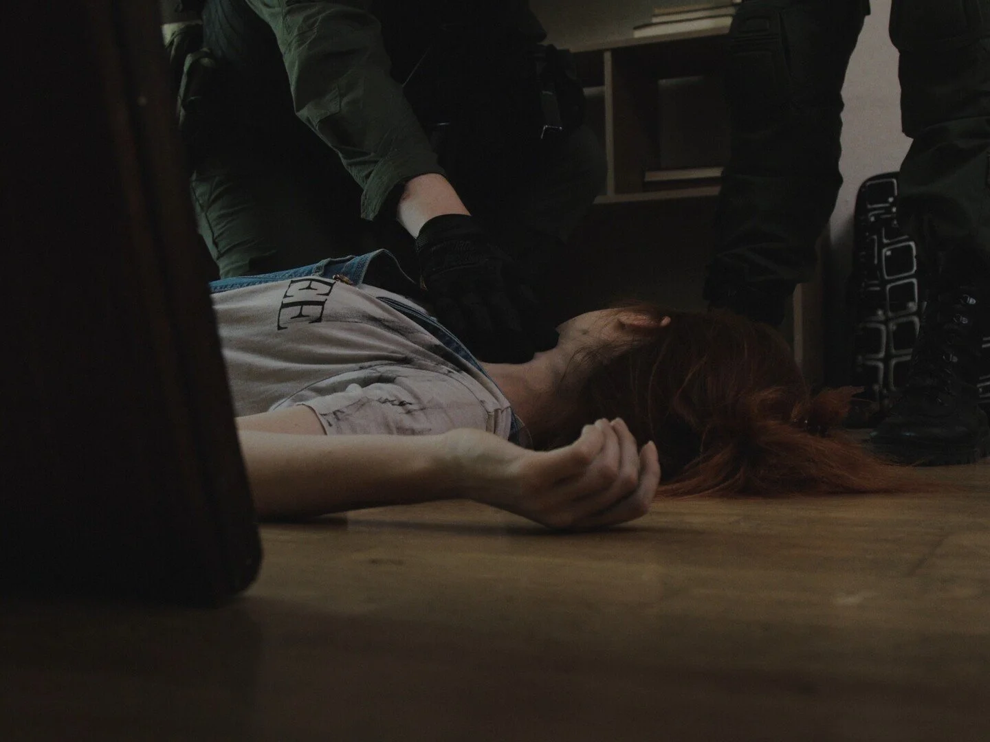 A woman with red hair lying on the floor with her eyes closed, surrounded by several people, some wearing black boots, appearing to assist or restrain her.