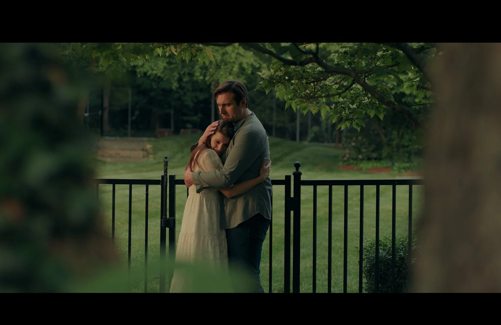 A man and a woman embracing each other emotionally outside on a grassy area, with trees and a fence in the background.
