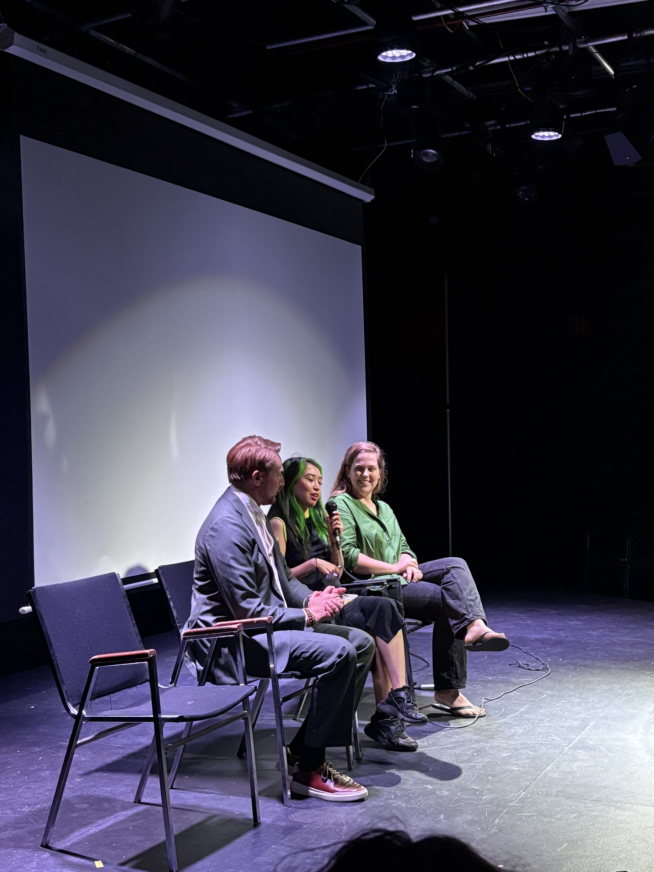 Three women sitting on stage chairs; the woman in the middle holds a microphone and talks, while the others listen.
