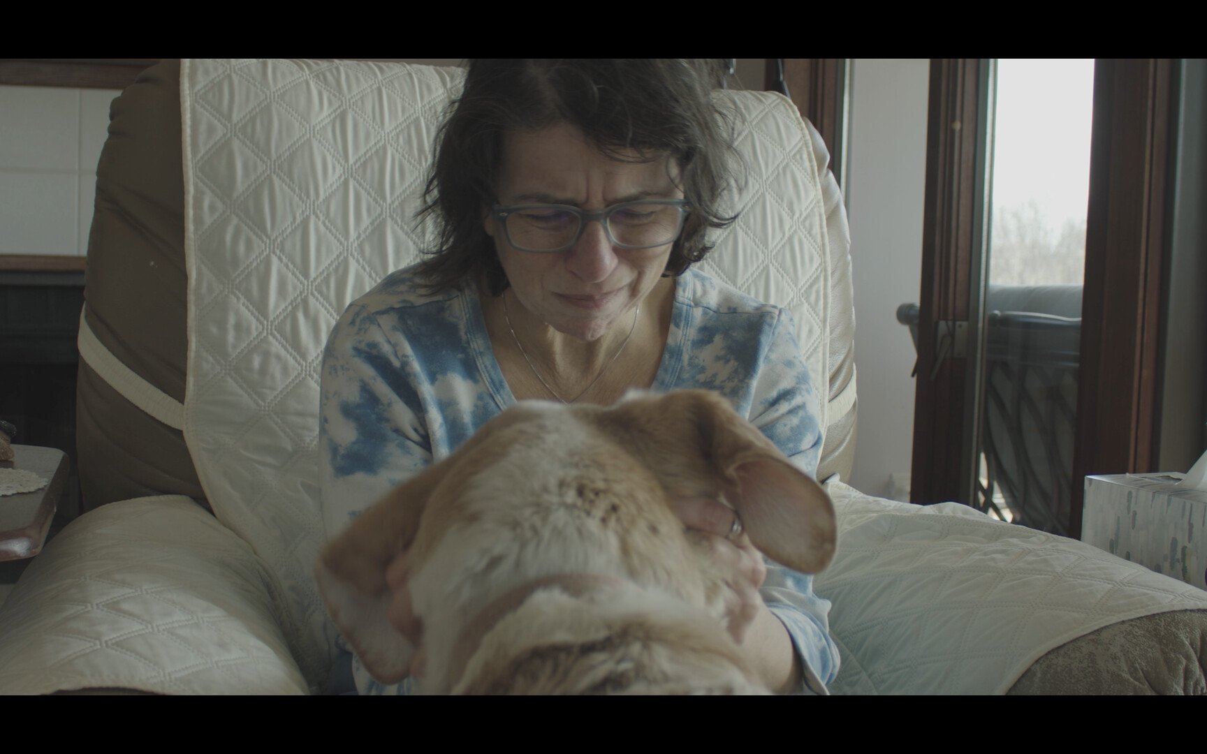 An older woman with glasses and curly hair, wearing a blue tie-dye shirt, sitting in a recliner, smiling and holding a large orange and white cat on her lap, in a cozy living room with natural light.