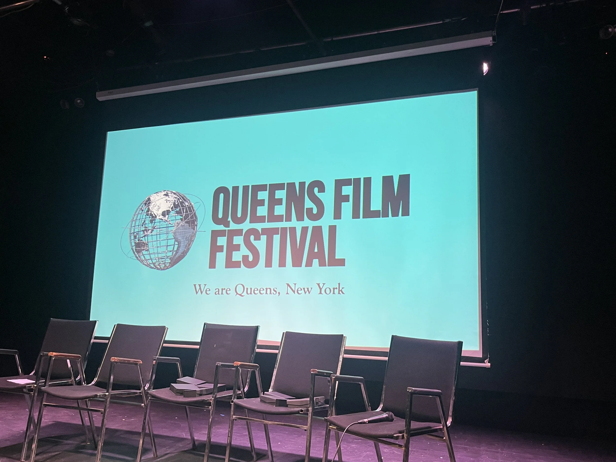 Stage with a large projection screen displaying 'Queens Film Festival' logo and text, with four chairs in front and a black background.