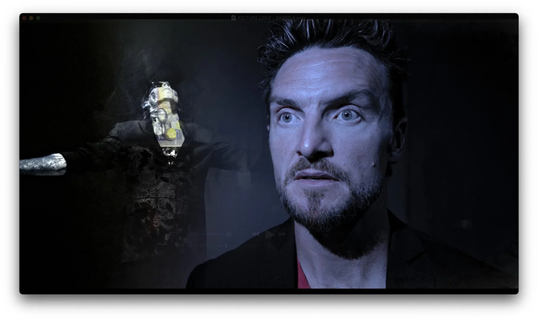 Close-up of a man with bluish eyes and a beard, looking serious, with a dark background and a blurry figure holding a gun in the background.