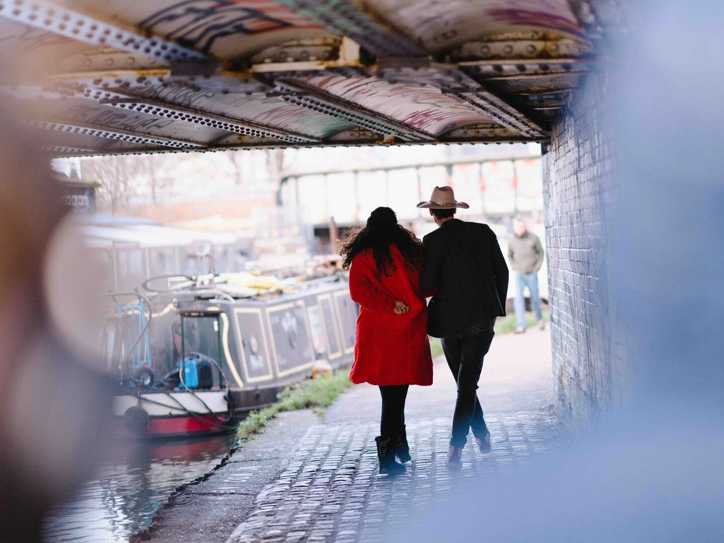 A couple walking together under a bridge near a canal, with a boat docked along the waterway and a man in the background.