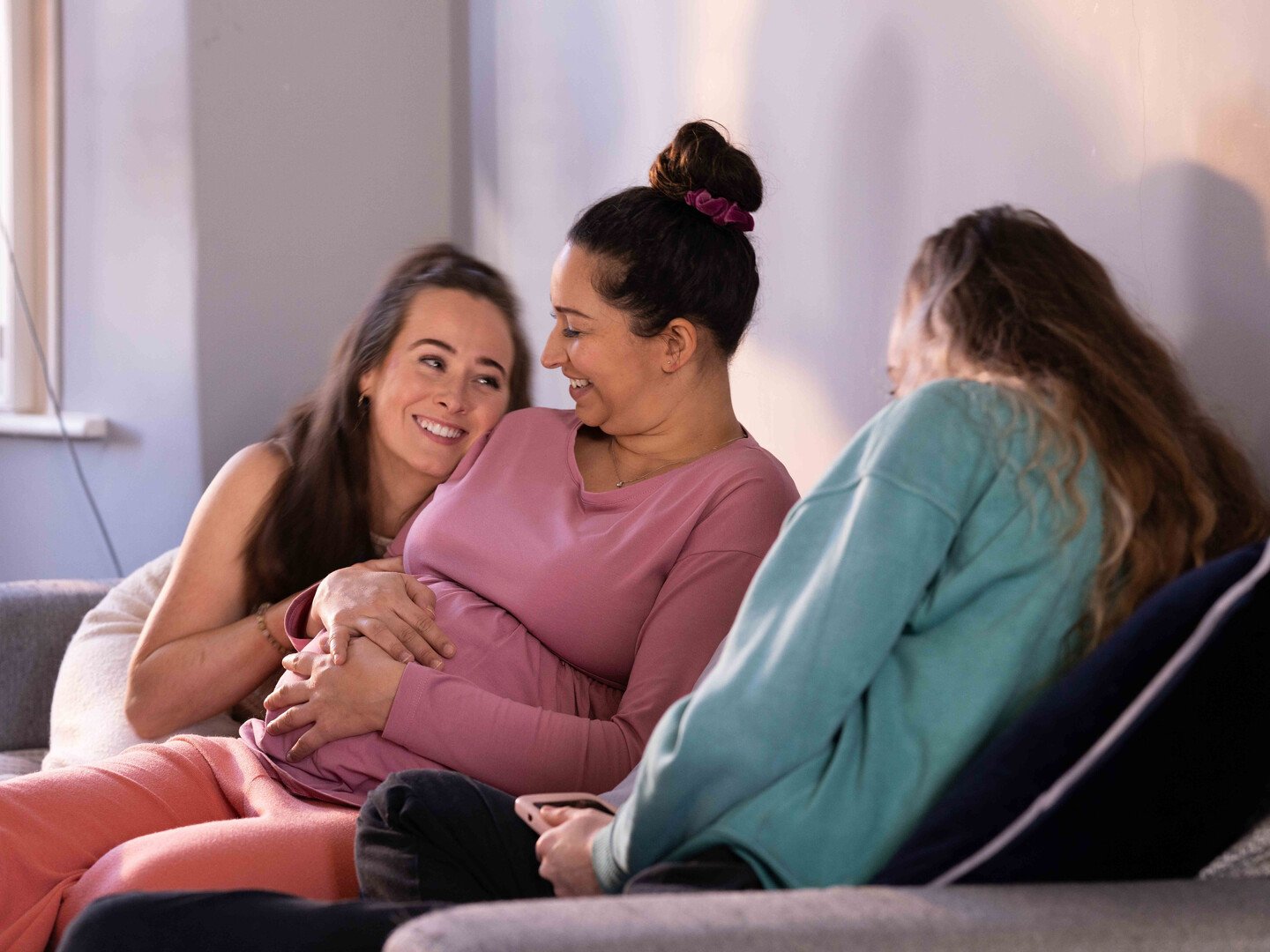 Three women sitting on a couch, smiling and sharing a joyful moment, one woman appears pregnant, in a cozy indoor setting.