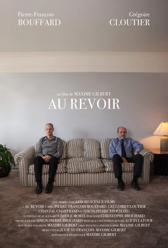 Two men sitting on a striped sofa in a living room. One is bald and wearing glasses, the other has a beard and is also wearing glasses. There is a lampshade on a side table to the right, and a potted plant on the left. The wall behind them has a poster for a film called "AU REVOIR" with names of actors and filmmakers written above and below.