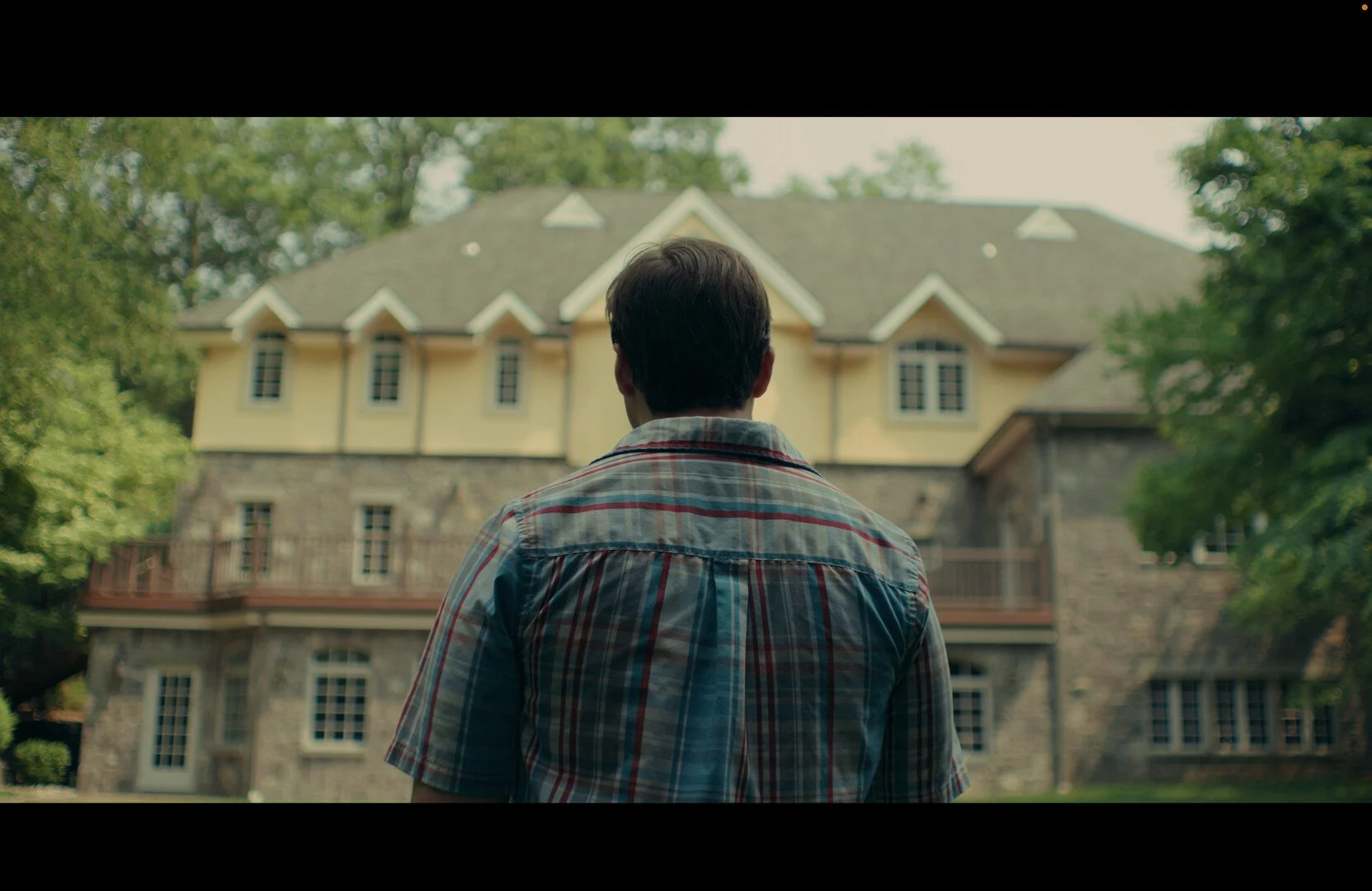 Man in plaid shirt standing in front of large house with stone and yellow exterior, lush green trees surrounding the property.
