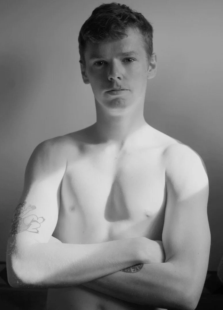 A black-and-white portrait of a young man with short hair, shirtless, crossing his arms, looking directly at the camera with a neutral expression, against a plain background.