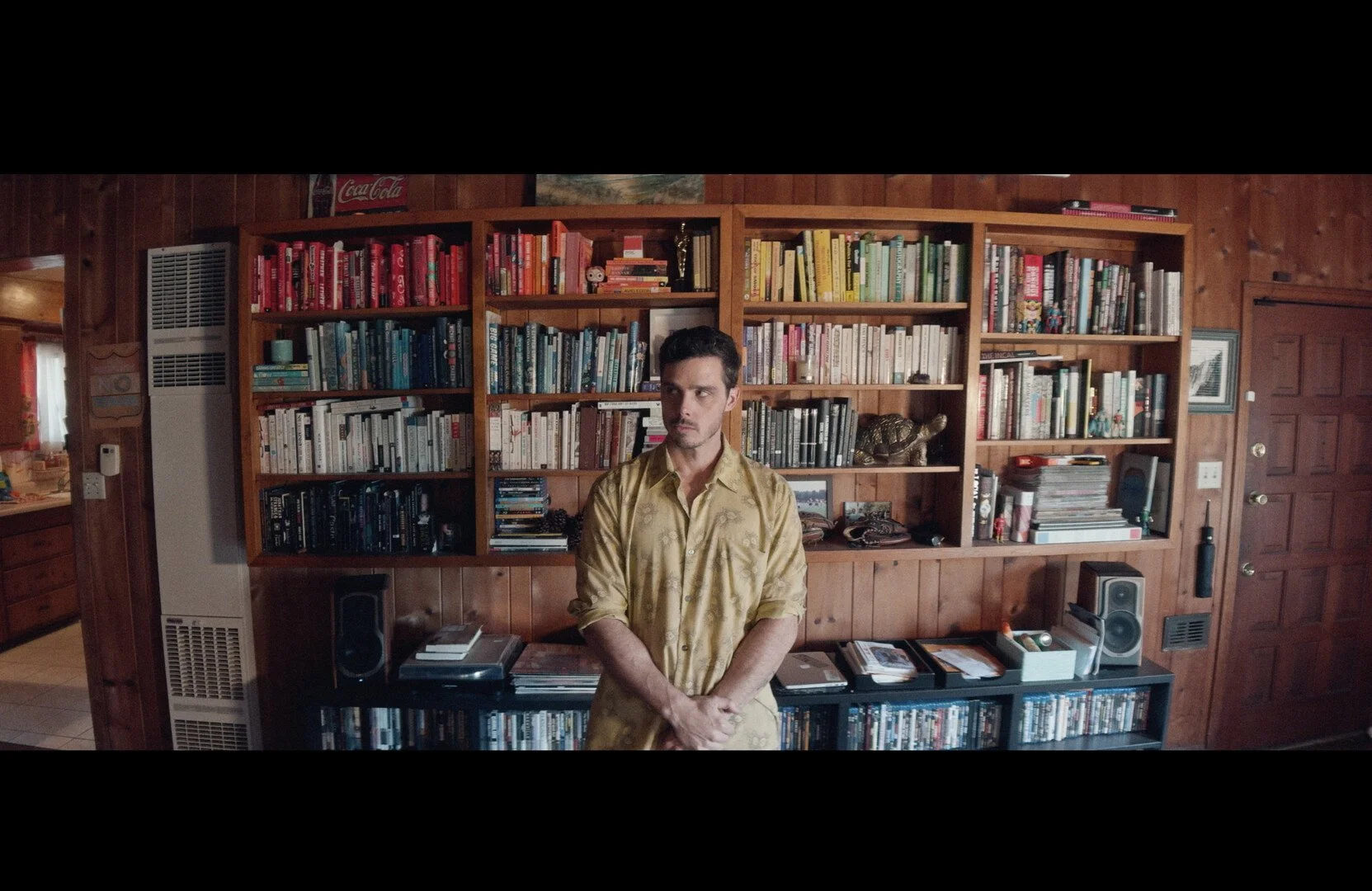 A man standing in front of a large wooden bookshelf filled with books, in a cozy wood-paneled room.