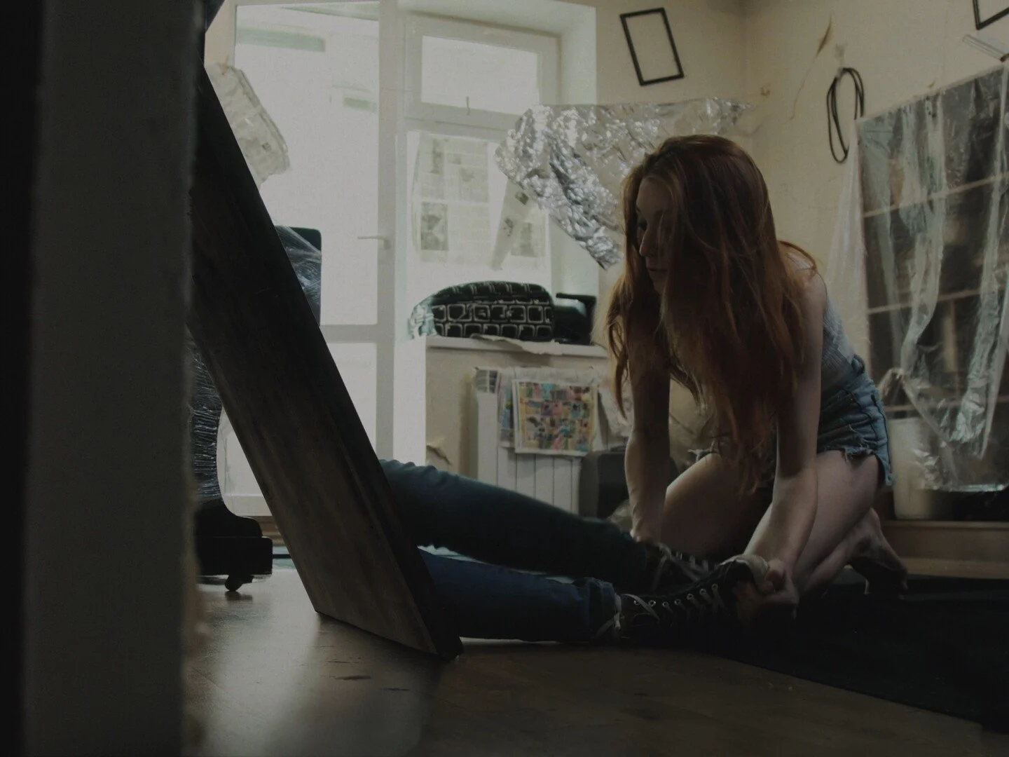 A young woman with red hair, wearing a plaid top and denim shorts, is kneeling on the floor. She is holding the foot of a person lying on the ground, whose face is not visible. The person has black pants and black shoes. The setting appears to be a cluttered room with large windows, some framed pictures on the wall, and various objects covered with plastic or foil for protection.